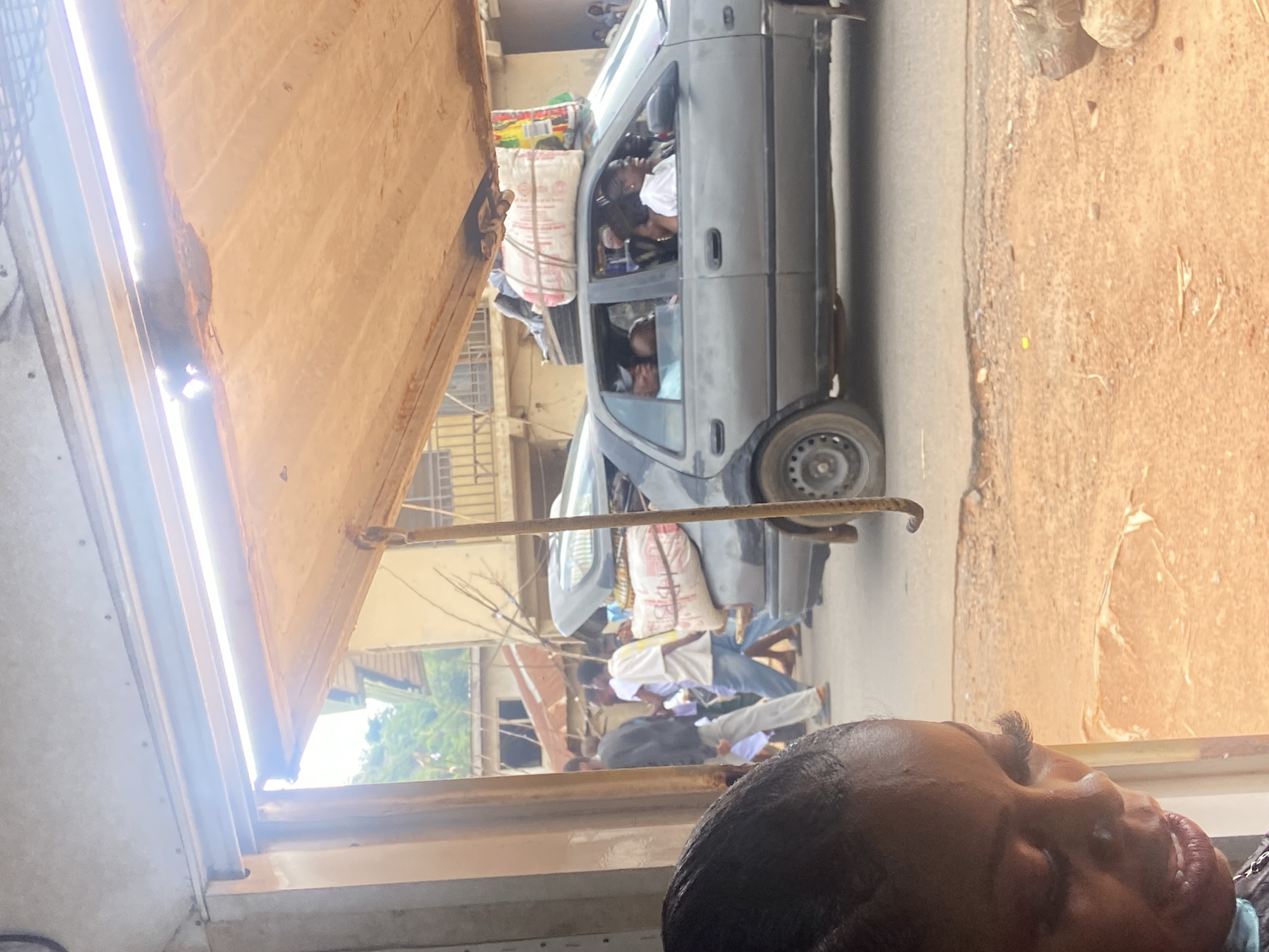 The image captures a scene in Magongo, Nigeria, viewed through a partially opened window of what appears to be a vehicle. A gray car, overloaded with people and goods on its roof, is parked on a street. Numerous people are standing or walking around the vehicle. The foreground includes a portion of a person's face and hair, presumably the photographer. The buildings in the background suggest an urban environment, and the visible light conditions indicate it is daytime.