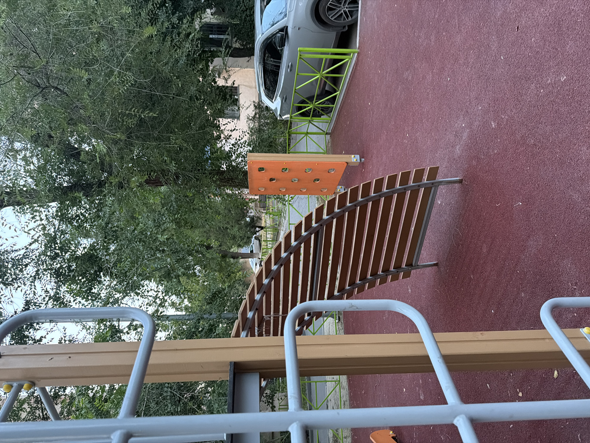 The image shows a section of an outdoor playground in Shymkent, Kazakhstan. In the foreground, there is a portion of a climbing structure with grey metal bars and a tan wooden post. Adjacent to this is a curved wooden structure with horizontal slats. To the right, a bright orange climbing wall with circular handholds stands near a light grey SUV parked behind a lime green metal fence. The ground is covered in a reddish-brown rubber safety surface. Behind the playground equipment, mature trees with green foliage create a natural backdrop, and a multi-story building is visible in the distance. The sky is overcast.