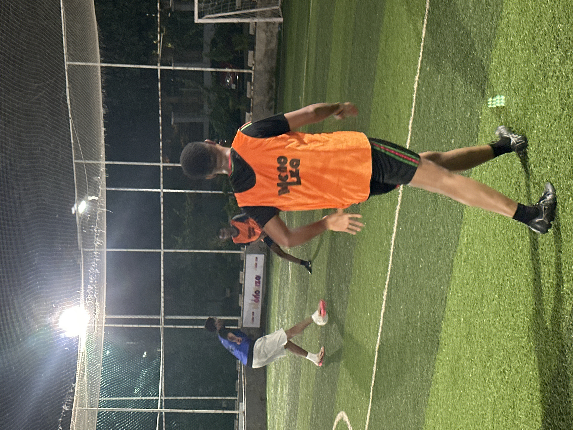 The image depicts a nighttime scene at an outdoor artificial turf soccer field in Abuja, Nigeria. Three individuals are visible on the field. In the foreground, a young man wearing an orange bib over a black t-shirt and black shorts with green and red stripes is walking away from the camera. He has dark skin and short black hair. To his left and further back, another young man is in motion, kicking a white soccer ball. He is wearing a blue sleeveless jersey, white shorts, white socks, and pink and white soccer cleats. His back is partially to the camera. In the background, a third young man, also wearing an orange bib over a dark shirt and dark shorts, is partially visible, appearing to be stationary. The field is marked with white lines. Above the field, a netting system encloses the area, with bright artificial lights illuminating the playing surface. The background beyond the netting shows dark vegetation and some structures. A white soccer goal is visible on the right side of the image. A sign with text, possibly a logo, is present on a barrier in the middle ground.