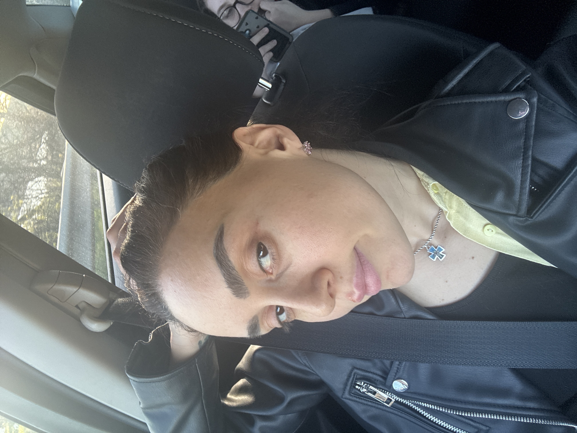 A person is seated in a car, wearing a black leather jacket, a black shirt, and a yellow collared shirt underneath. They are also wearing a silver necklace with a cross pendant. A seatbelt is fastened across their chest. The person has dark hair pulled back, fair skin, and light eye color. Another person is visible in the background, holding a smartphone. The scene appears to be inside a car, possibly moving or parked. It is not possible to determine whether this image was taken in Tbilisi, Georgia from the given information.