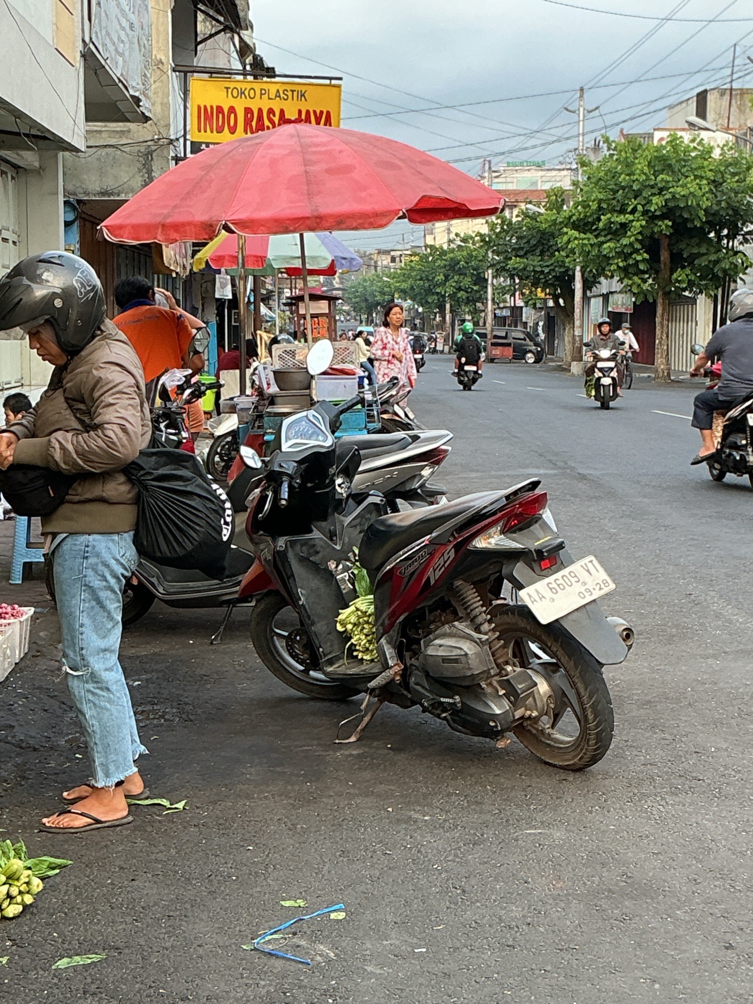 The image depicts a street scene in Magelang, Indonesia, characterized by moderate pedestrian and vehicular traffic under an overcast sky. In the foreground, a person wearing a brown jacket, blue jeans, and a helmet stands next to a motorcycle. To their left, a crate of red onions is visible. Several motorcycles are parked and in motion on the street, including one with the license plate "AA 6609 VT" which expires in September 2028. On the right side of the street, there are shops and trees. A prominent yellow sign reads "TOKO PLASTIK INDO RASA JAYA". Red umbrellas are set up, suggesting a market or roadside stalls. People are seen walking and riding motorcycles in the background. The overall environment appears to be a typical urban street with commercial activity.