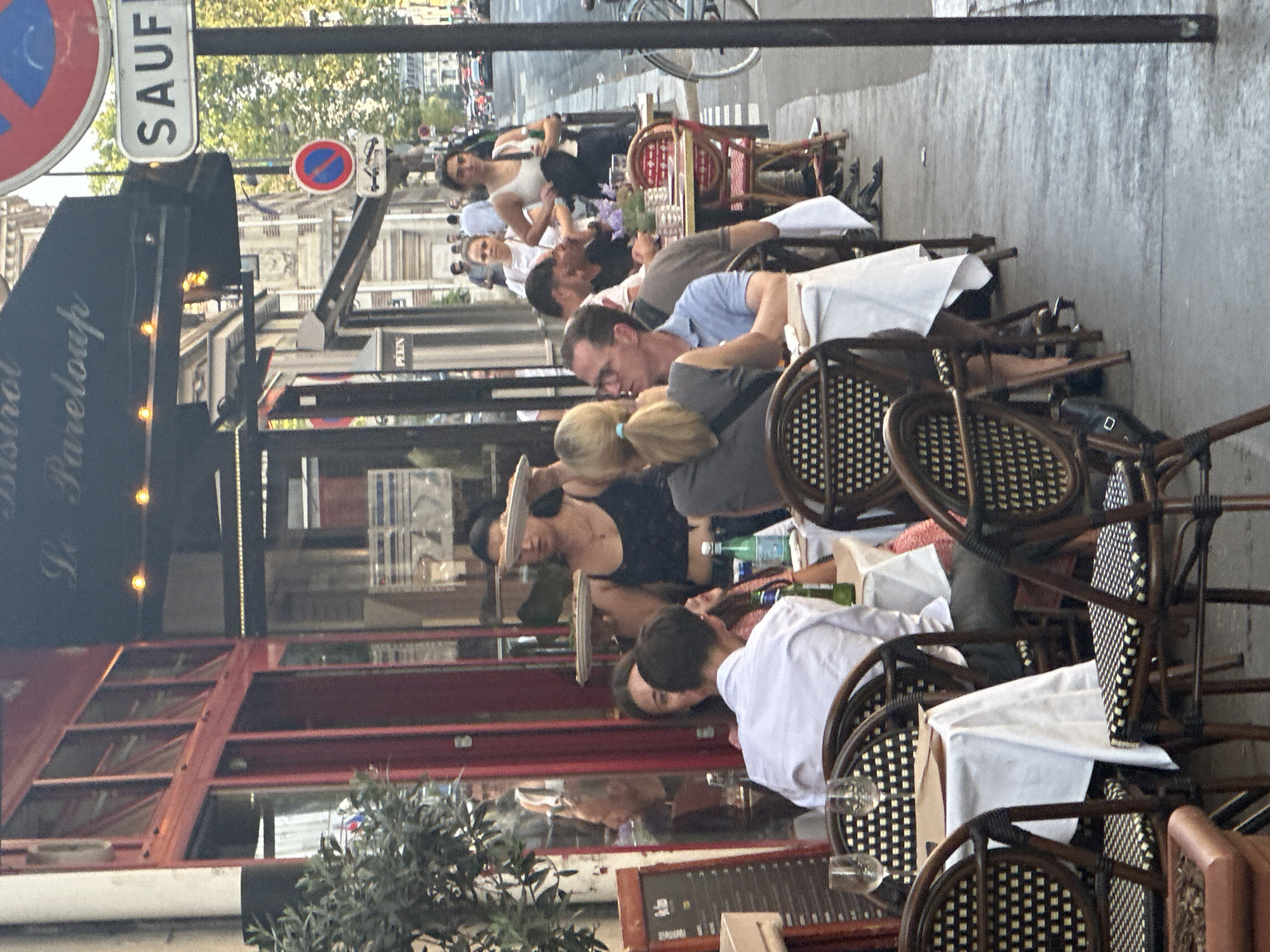 The image depicts an outdoor seating area of a Parisian bistro, identified by the awning text "Bistrot Pareloupa". Several tables are set with white tablecloths and chairs, some occupied by diners. In the foreground, a man wearing a white shirt and glasses is seated at a table. To his right, a woman with blonde hair pulled back is also seated. Further back, a waiter or server is visible holding plates. Behind the outdoor seating, a red-fronted building with large windows lines the street. A sidewalk is present, with pedestrians walking in the background. Traffic signs, including a "SAUF" sign, are visible on a pole, indicating a street scene in Paris. The overall atmosphere suggests a typical Parisian dining experience on a street-side cafe.