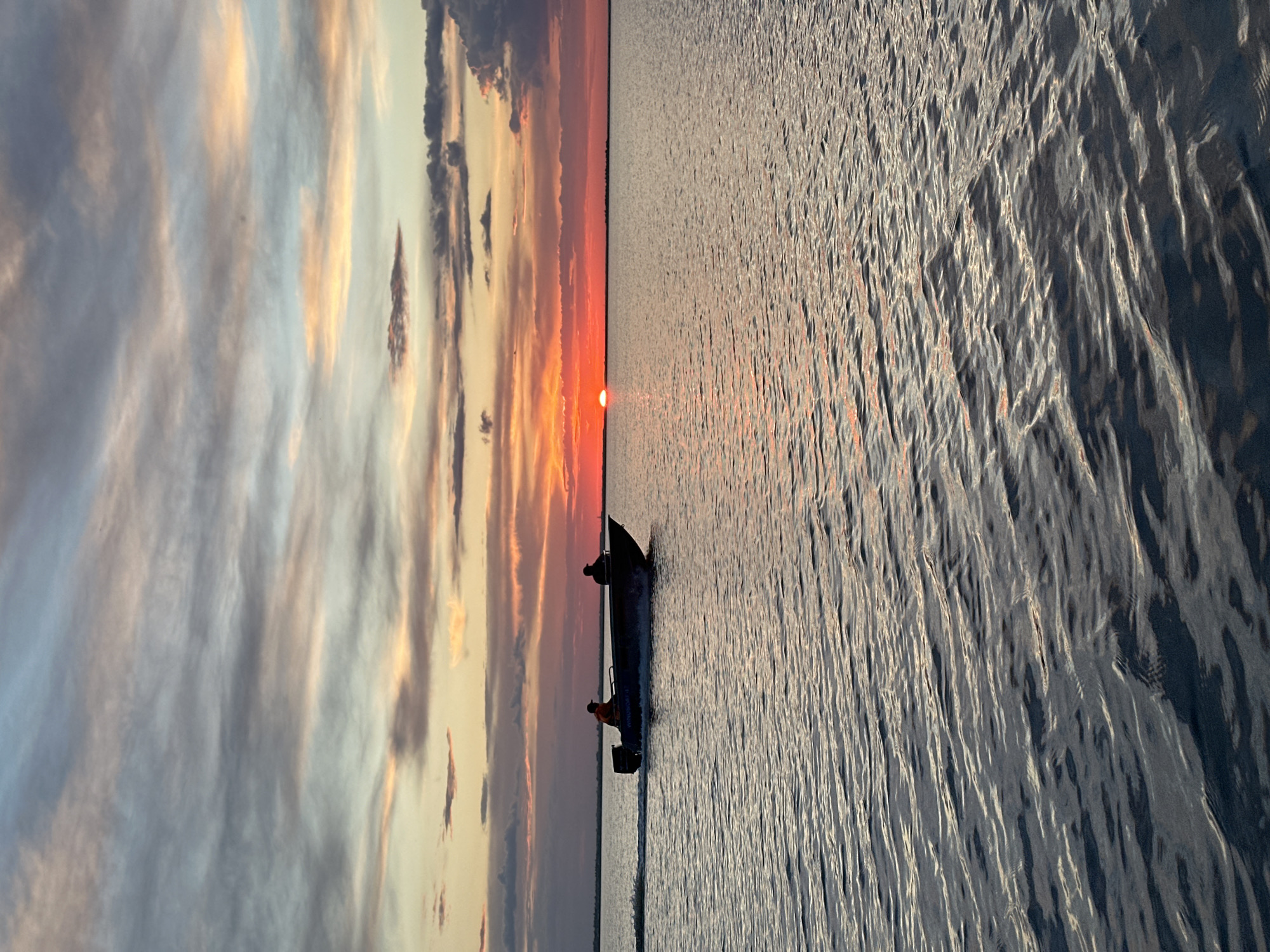A motorboat with two silhouetted individuals on board travels across a body of water at sunset. The sun, a bright orange orb, is partially visible on the horizon, casting a warm glow across the sky. The sky is filled with a mix of wispy clouds, tinged with shades of pink, orange, and gray. The water's surface is rippled, reflecting the colors of the sunset. The scene suggests a tranquil evening, possibly on a lake or river. The location is the city of Kokshetau, Kazakhstan.