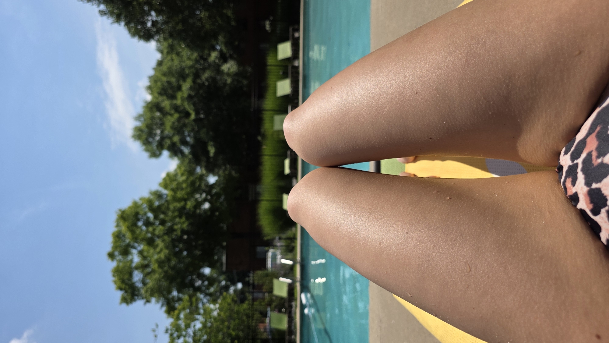 A first-person perspective shot shows two sun-tanned legs in the foreground, lying on a yellow lounge chair. The legs are wet with small water droplets visible on the skin, suggesting a recent dip in the water. A leopard-print bikini bottom peeks out from between the legs. The background reveals a bright blue swimming pool with reflections of the sun. Beyond the pool, lounge chairs are neatly arranged on a concrete deck. Lush green trees and a brick building are visible further back, under a clear blue sky with a few wispy clouds. The scene evokes a leisurely summer day at an outdoor pool.