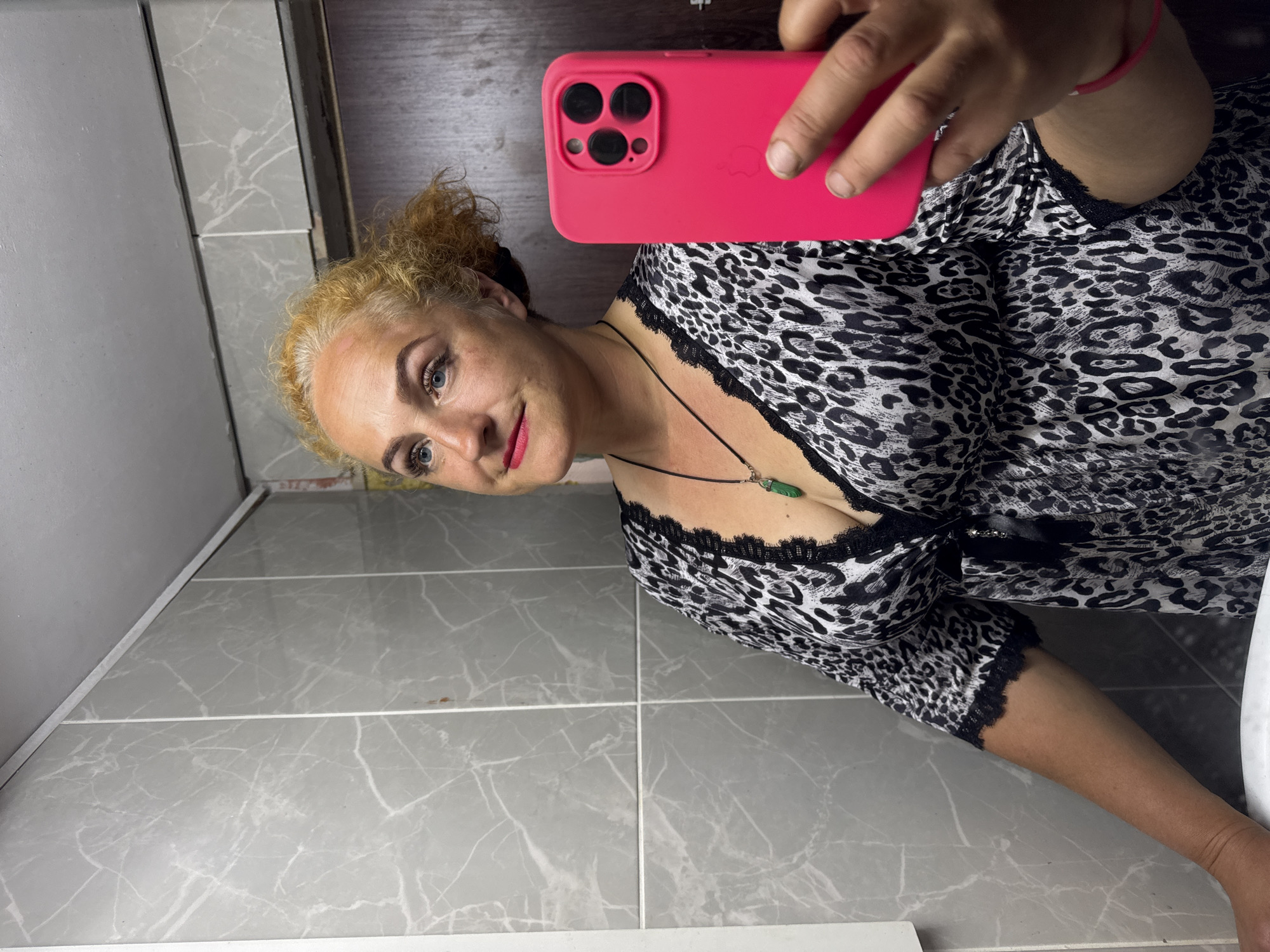 The image shows a woman taking a mirror selfie in a bathroom. She has blonde, curly hair and is wearing a black and white leopard print dress with black lace trim. She is wearing a necklace with a green pendant. She is holding a bright pink phone in her right hand, which is pointed towards the mirror. The bathroom has grey marble-patterned tiles on the wall and a white sink. The lighting is dim, and there are some shadows visible. The location is identified as Chaikovsky, Russia.