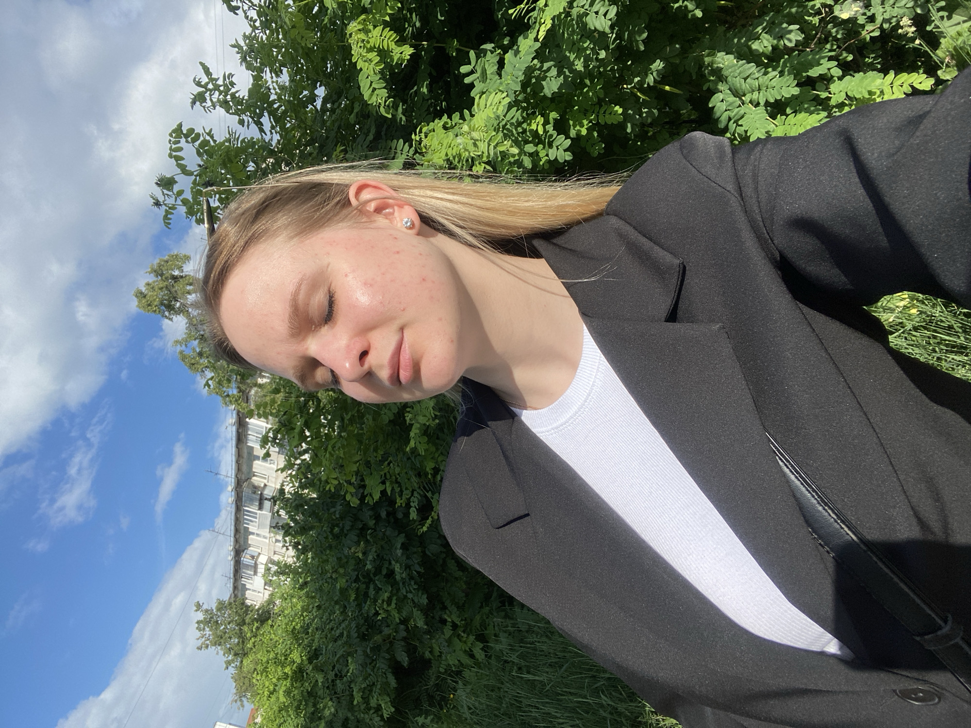 The image shows a young woman with blonde hair and fair skin, wearing a black blazer over a white top. She has her eyes closed and appears to be enjoying the sun. She is wearing a small diamond stud earring in her left ear. In the background, there are lush green trees and a building with balconies, suggesting an urban setting. The sky above is blue with scattered white clouds. Based on the architectural style of the building and the surrounding vegetation, the location is likely an outdoor area in the city of Chaikovsky, Russia, possibly a residential district.