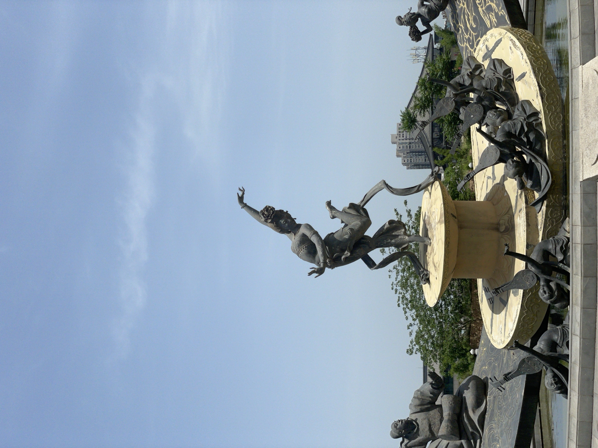 The image depicts a large bronze statue in Lintong District, China. The statue features a woman with her arm raised and a man beside her, possibly a representation of Chang'e, a Chinese moon goddess, and her husband. The figures are positioned on top of a cylindrical structure adorned with other smaller sculptures of people playing musical instruments. The background includes buildings and trees, indicating an urban or park-like setting. The sky is mostly clear with a blue hue.
