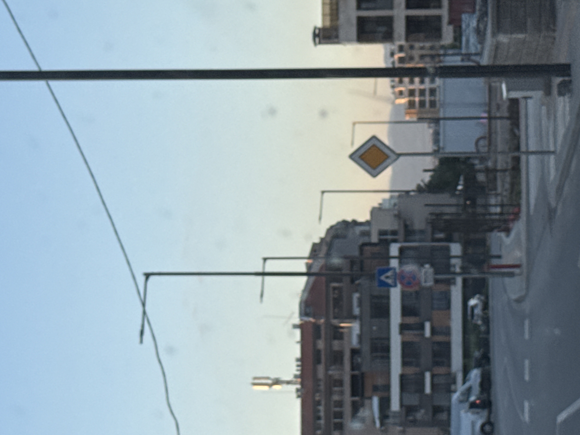 The image shows an urban scene in Sofia, Bulgaria. A yellow diamond-shaped "priority road" sign is visible. A road sign indicating "give way" is also present. Buildings are in the background. A street with at least one vehicle on it runs vertically on the right. A street lamp is present on the left. There are overhead wires in the image.