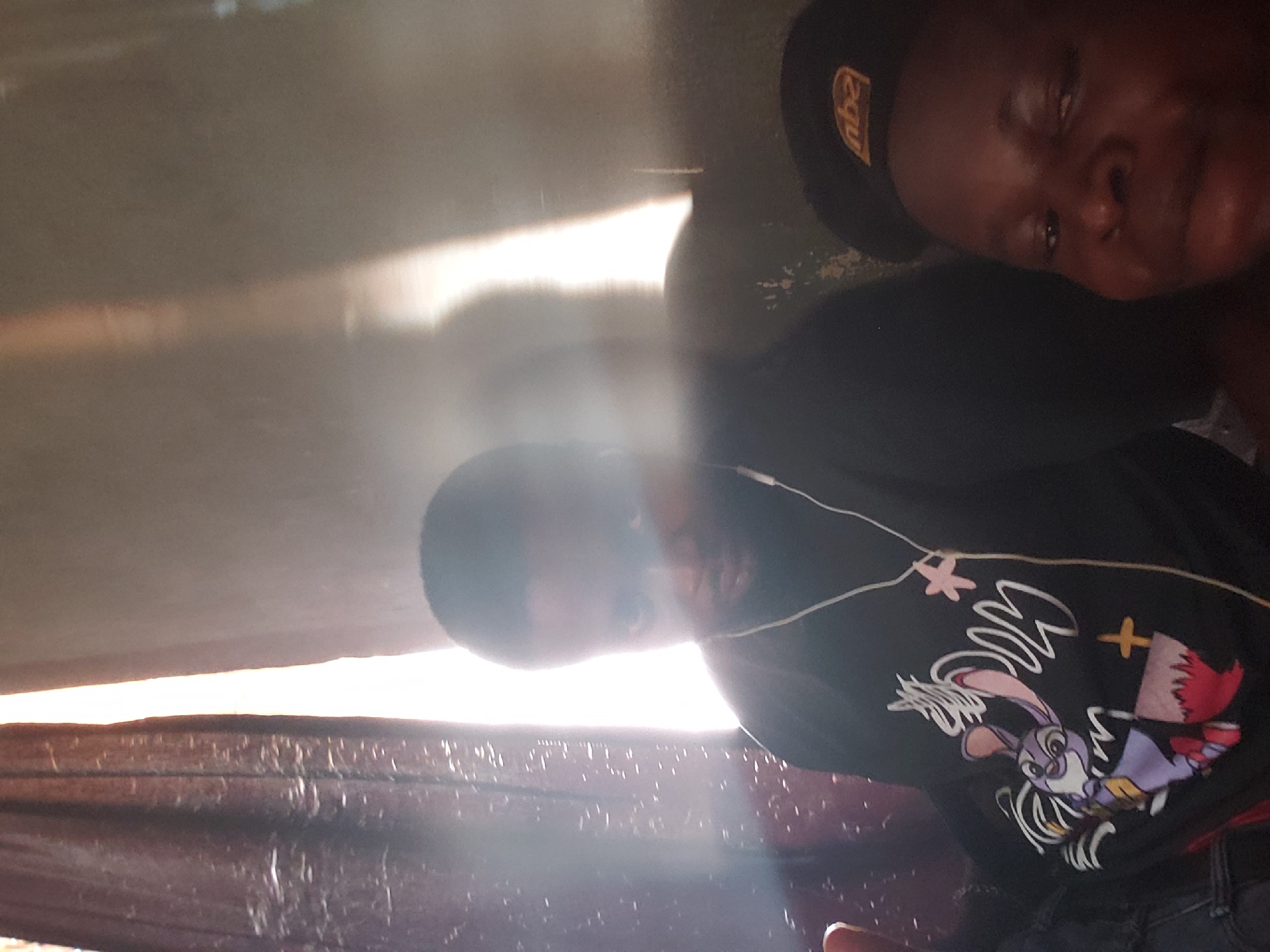 The image, when correctly oriented, is a portrait-style photograph taken indoors, showcasing two individuals in a dimly lit environment dramatically illuminated by a bright horizontal light source.
The individual in the foreground, positioned on the right side of the frame, is a person with dark skin. They are wearing a black cap adorned with a yellow "NBA" logo. Their face, seen from the chin upwards, appears to have a calm, perhaps relaxed or pensive expression, with their gaze directed slightly away from the camera. They are dressed in a dark-colored top.
The second individual is located further back, on the left side of the frame, partially obscured by the intense light. This person, also with dark skin, is wearing a black t-shirt featuring a colorful cartoon character – a purple, rabbit-like figure, possibly Bugs Bunny, depicted with a red cape and dynamic white motion lines. White wired earbuds are visible in their ears.
A prominent and intense beam of light cuts horizontally across the image, suggesting natural light streaming through a narrow opening or window. This strong illumination highlights numerous dust particles in the air, creating visible light rays and a slightly hazy atmosphere. The background and surrounding areas are dark and indistinct, appearing to be dark brown or black surfaces, possibly walls or fabric. A textured, dark surface resembling upholstery or a partition is visible in the lower part of the image.
Both individuals appear to be in a relaxed or resting posture, with the person wearing earbuds potentially listening to audio. The strong natural light suggests the photograph was taken during the daytime. The only visible text is the "NBA" logo on the cap.