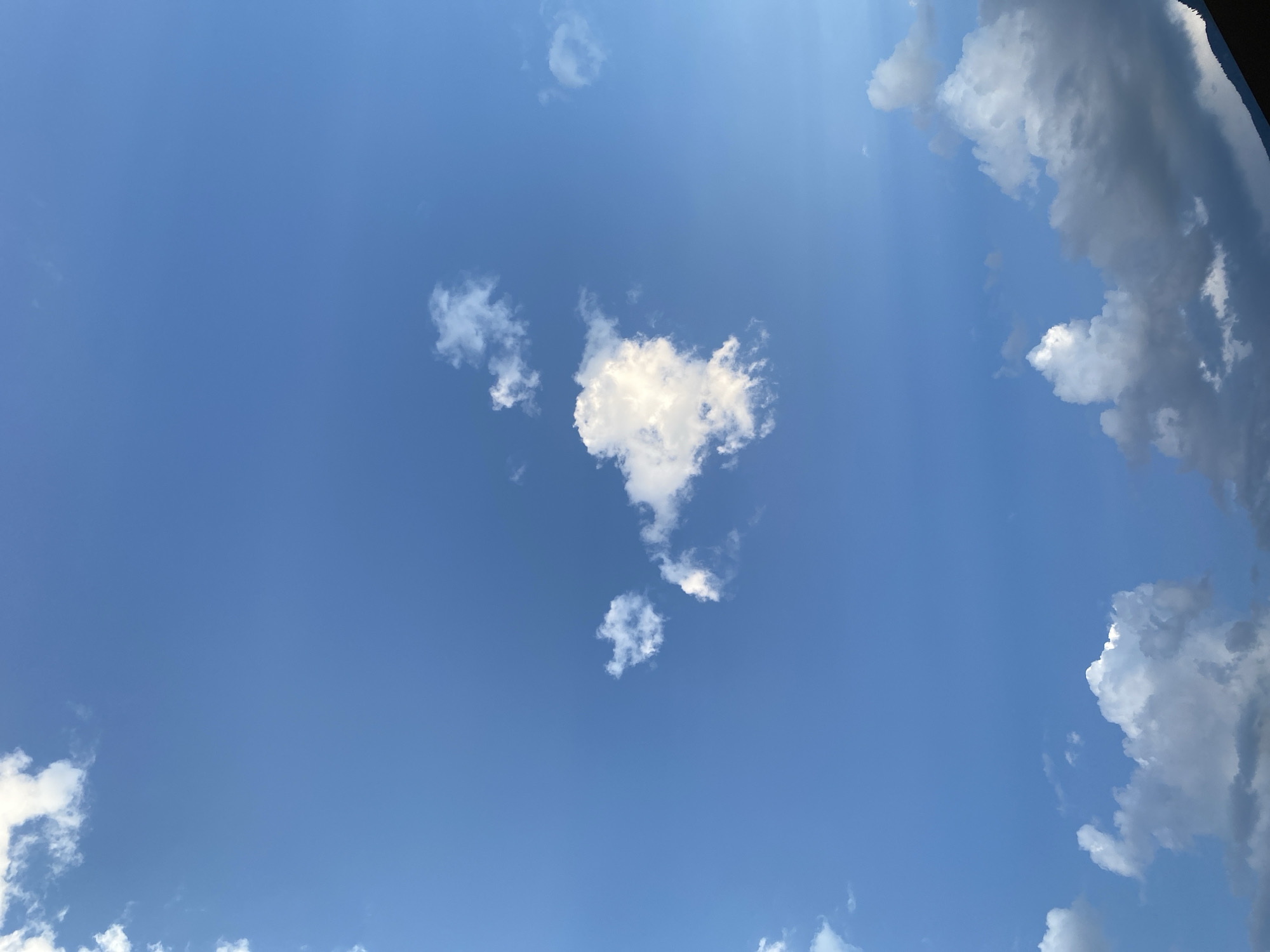 The image depicts a clear blue sky with scattered white cumulus clouds. The clouds are mostly fluffy and detached, with some appearing larger and more substantial than others. A few wispy clouds are also visible. The lower portion of the image shows a dark, silhouetted mountain range, suggesting a natural landscape setting. The overall scene is serene and peaceful. The location is the city of Meran - Merano, Italy.
