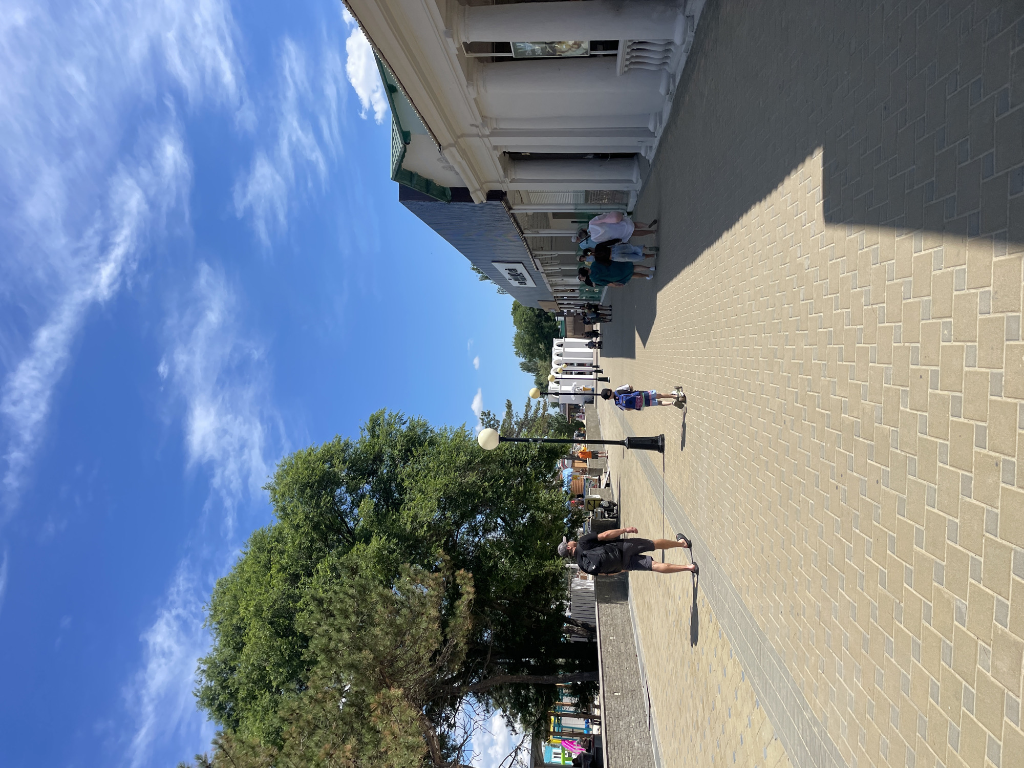 The image shows a sunny day on a pedestrian walkway in an urban environment, possibly a resort town. The walkway is paved with light-colored bricks arranged in a diamond pattern. On the left side of the walkway, there is a low stone wall with a row of trees behind it, including a large, mature deciduous tree and several evergreen trees. Beyond the trees, there appear to be some colorful structures, possibly kiosks or children's play areas. On the right side of the walkway is a row of white buildings with classical architectural elements, including large columns and ornate railings. Some of these buildings have arched entrances. Further down the walkway, a darker, more modern building with a dark facade is visible.
Several people are walking along the walkway. In the foreground, a man wearing a black t-shirt, grey shorts, a grey cap, and black sandals with a black backpack is walking away from the camera. To his right, a young boy is riding a scooter, also walking away from the camera, and is wearing a blue backpack. Further ahead on the right side of the walkway, a group of people are walking together. In the background, more people can be seen walking, and there are some light poles along the walkway. The sky is a clear blue with scattered white clouds. The scene suggests a lively public space on a pleasant day.