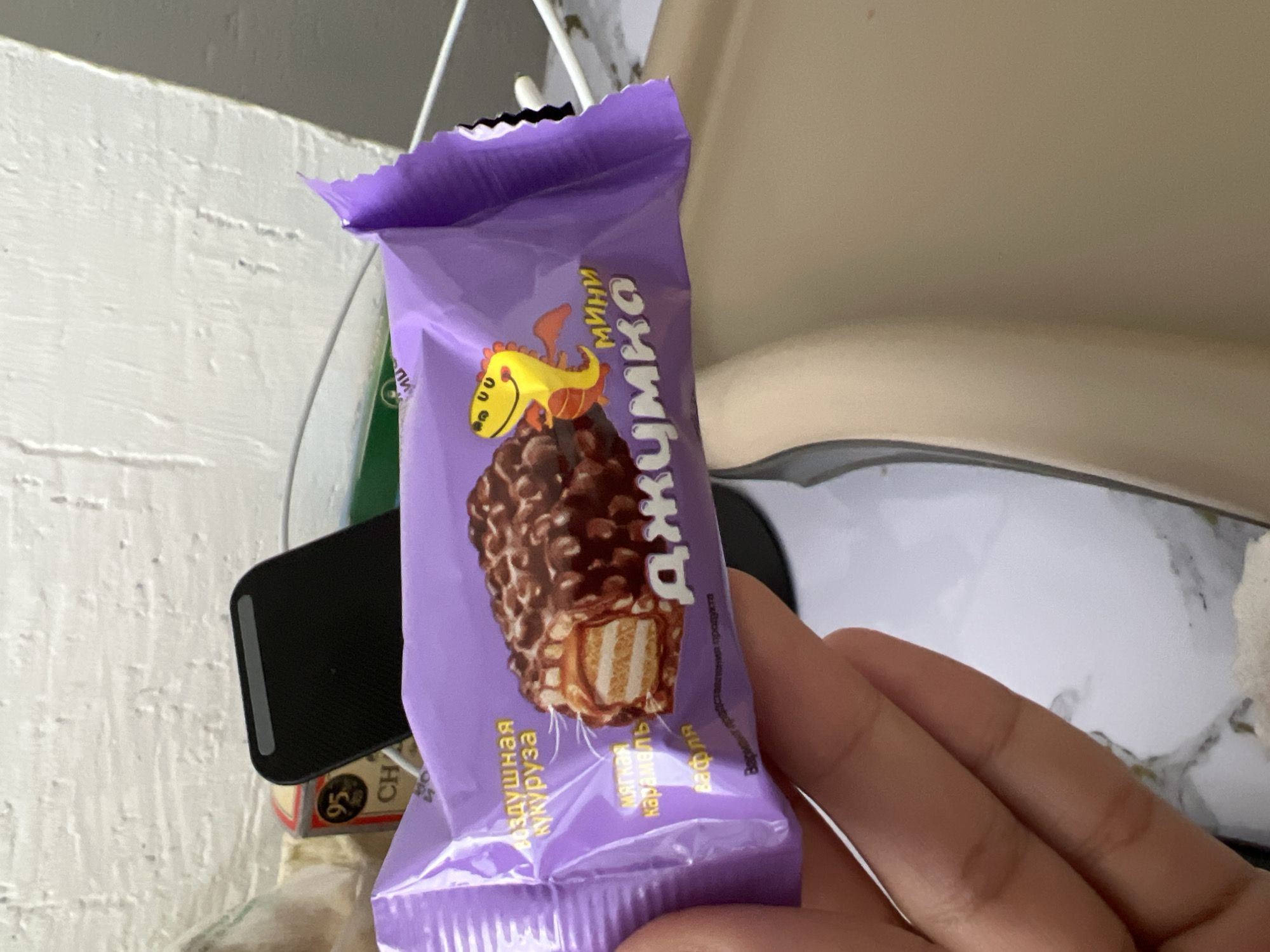The image shows a hand holding a purple candy bar wrapper with a picture of a chocolate-covered snack. The wrapper has text in Cyrillic script. The background is blurred and shows a white textured wall and some objects that are not clearly identifiable. There is no information in the image that suggests the location is Tokmok City, Kyrgyzstan.