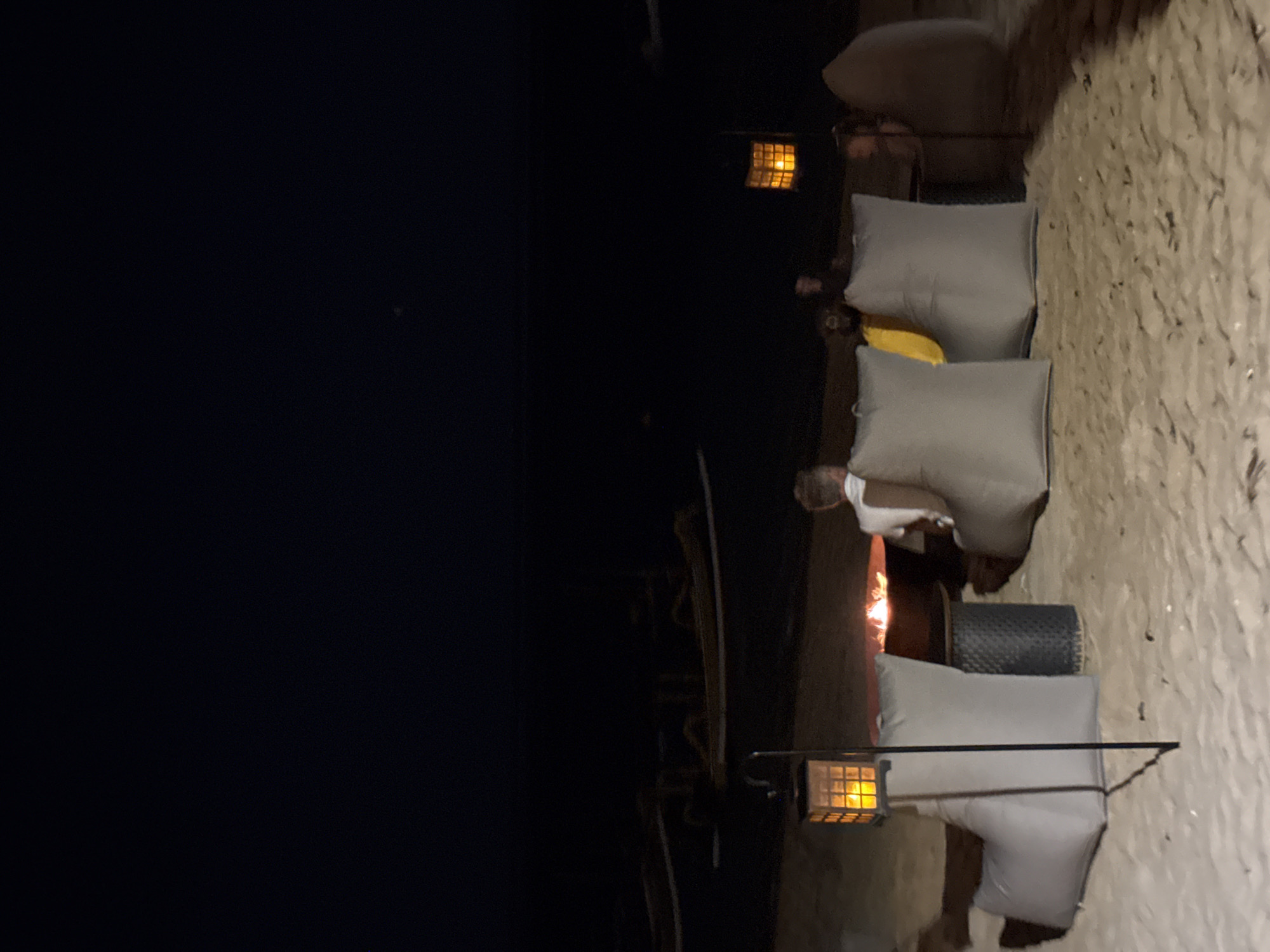 The image depicts a nighttime scene on a sandy beach, presumably in Denpasar, Indonesia. In the foreground, there are several large, light gray beanbag chairs arranged around a fire pit. A dark, cylindrical table is positioned between some of the beanbags and the fire pit. A metal pole with a square lantern, illuminated with a warm yellow light, stands to the left of the beanbags. The fire pit contains a visible flame.
Further back, two individuals are seated in beanbag chairs. One person, viewed from the back, is wearing a white shirt and sitting in a beanbag chair to the right of the fire pit. Another person, wearing a yellow garment, is partially visible between two gray beanbag chairs, also facing away from the camera. A third, darker figure is seated behind them. In the background, the dark ocean meets a black sky. Several outrigger canoes, with their distinctive hulls and outriggers visible, are pulled up onto the beach or floating in the shallow water. The overall atmosphere is relaxed and tranquil, illuminated by the fire and the lanterns.