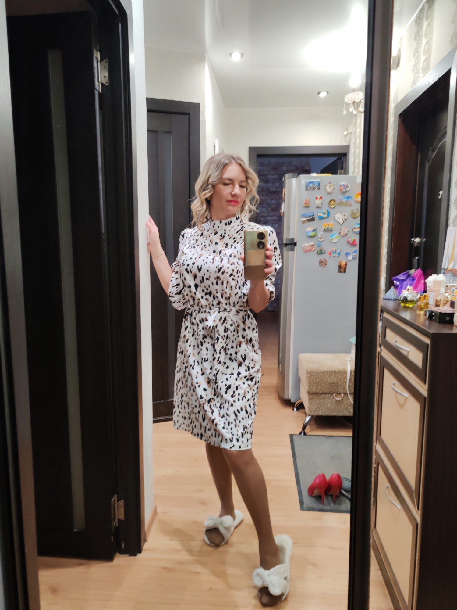 A woman stands in a hallway, posing for a mirror selfie. She is wearing a patterned dress, tights, and fuzzy slippers. The hallway has wood floors, dark wood doors, and a light-colored wall. A refrigerator covered in magnets is visible in the background. A cabinet sits to the right of the mirror with shoes next to it. The scene is set in an apartment in Petropavlovsk-Kamchatsky, Russia.