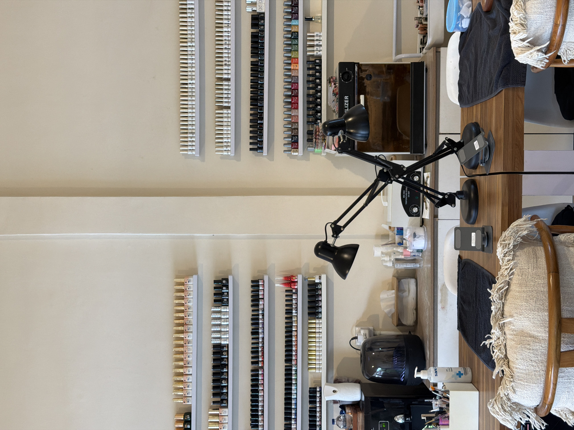 The image depicts a well-organized nail salon, likely in North Kuta, Indonesia. The primary focus is on the shelves lining the walls, which are filled with rows of nail polish bottles. These bottles are predominantly in shades of gold, silver, black, and various colors. The salon appears to be set up for multiple workstations, with a wooden table in the foreground featuring chairs and various nail care supplies.
On the table, there are two adjustable desk lamps, one black and one silver, positioned over the workspace. Several black towels are neatly folded on the table, and a hand sanitizer dispenser is visible. Also on the table are containers for nail tools and cotton pads. In the background, a sterilization unit is present, along with a dispenser for tissues. The overall environment is clean and professional, suggesting a busy and operational nail salon. There are no people visible in the image.