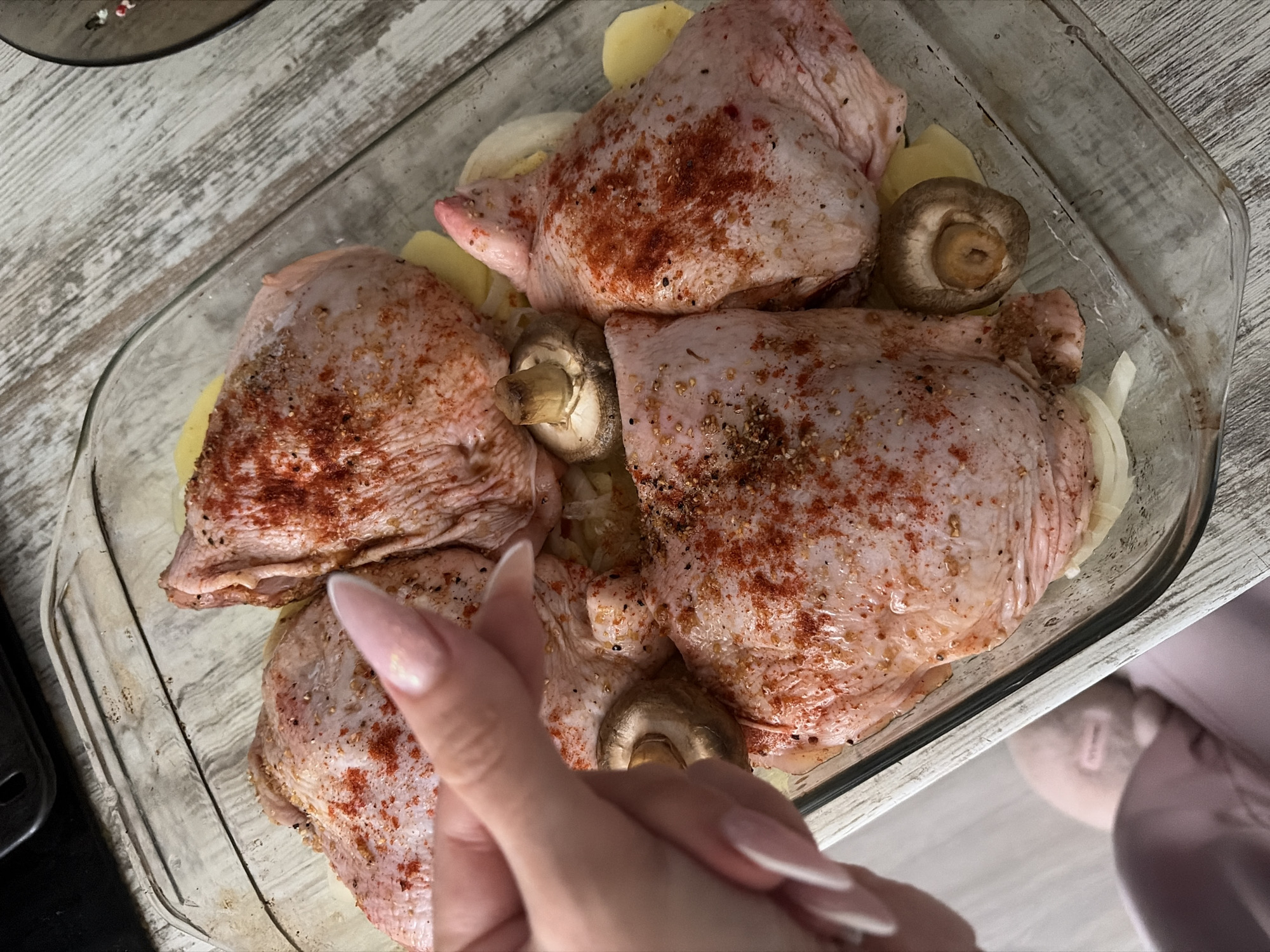 A top-down, close-up shot shows a clear glass baking dish filled with seasoned raw chicken pieces, potatoes, onions, and mushrooms. A hand with long, manicured nails is positioned in the foreground. The food is liberally seasoned with paprika and other spices. The background is a light-colored wood-grain surface. The setting suggests a home kitchen. It is unclear if this image was taken in the city of Saint Petersburg, Russia.