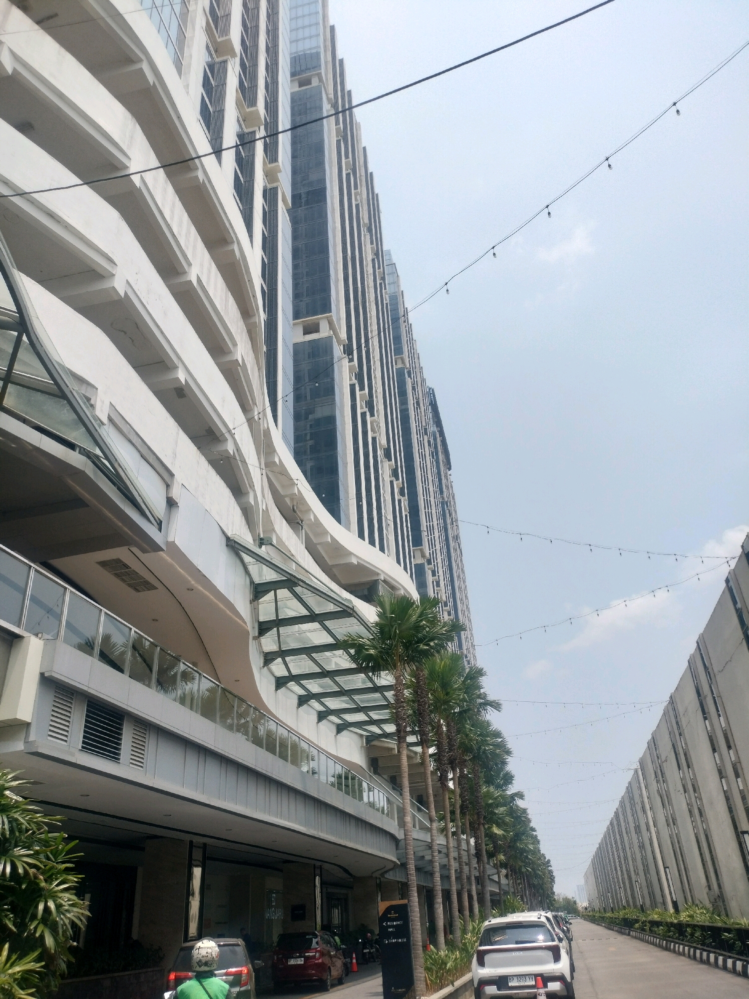 The image depicts a street scene in Batam, Indonesia, characterized by modern architecture and a clear sky. On the left, a towering multi-story building with a curved facade dominates the frame. The building features a combination of white, horizontal architectural elements and dark, reflective windows. Below the upper floors, a covered walkway with a glass roof is visible, lined with palm trees.
On the right side of the street, a long, textured concrete wall with vertical accents extends into the distance. Two parallel lines of string lights with bulbs are suspended in the sky above the street.
Several vehicles are present on the paved road. In the foreground, a white Kia SUV is parked facing away from the camera. To its left, a dark red car is also visible. A person on a green motorcycle wearing a white helmet is in the process of maneuvering through the vehicles. Further down the road, other vehicles and a person walking can be seen. The overall environment suggests a commercial or residential area within the city.