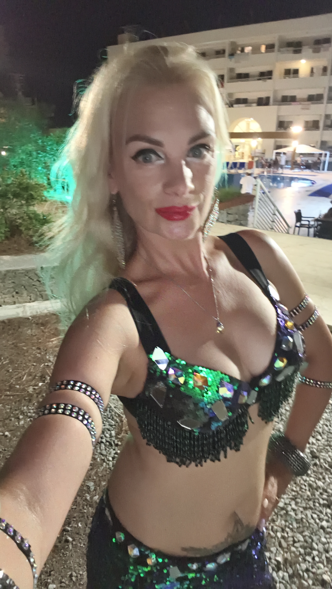 A woman with blonde hair and red lipstick is posing for a selfie in front of a brightly lit building at night. She is wearing a sequined belly dancing costume with a green and black bra and a dark skirt. She has large earrings and bracelets on her arms. In the background, there is a swimming pool and people sitting at tables. The location appears to be an outdoor venue in Kuşadası, Turkey.