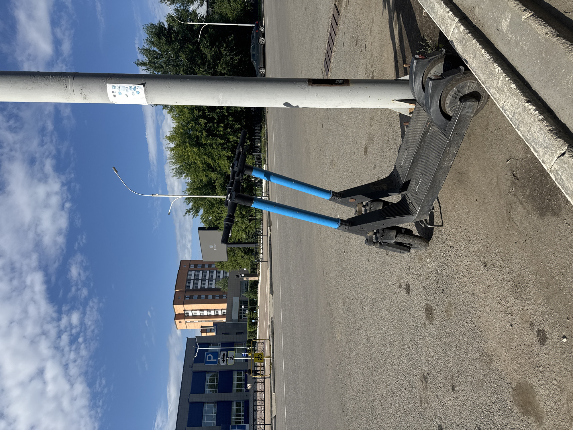 An electric scooter is parked next to a grey utility pole on a paved surface, possibly a sidewalk or parking area, adjacent to a street. The scooter is black with blue handlebars and appears to be a shared rental type. The utility pole has a white notice with several small icons and text attached to it. In the background, there are several buildings, including a blue and grey modern structure on the left and a reddish-orange multi-story building further back. Across the street, there is a dark-colored sedan. Trees line the background, and the sky is blue with scattered white clouds. The scene is set in Kokshetau, Kazakhstan.