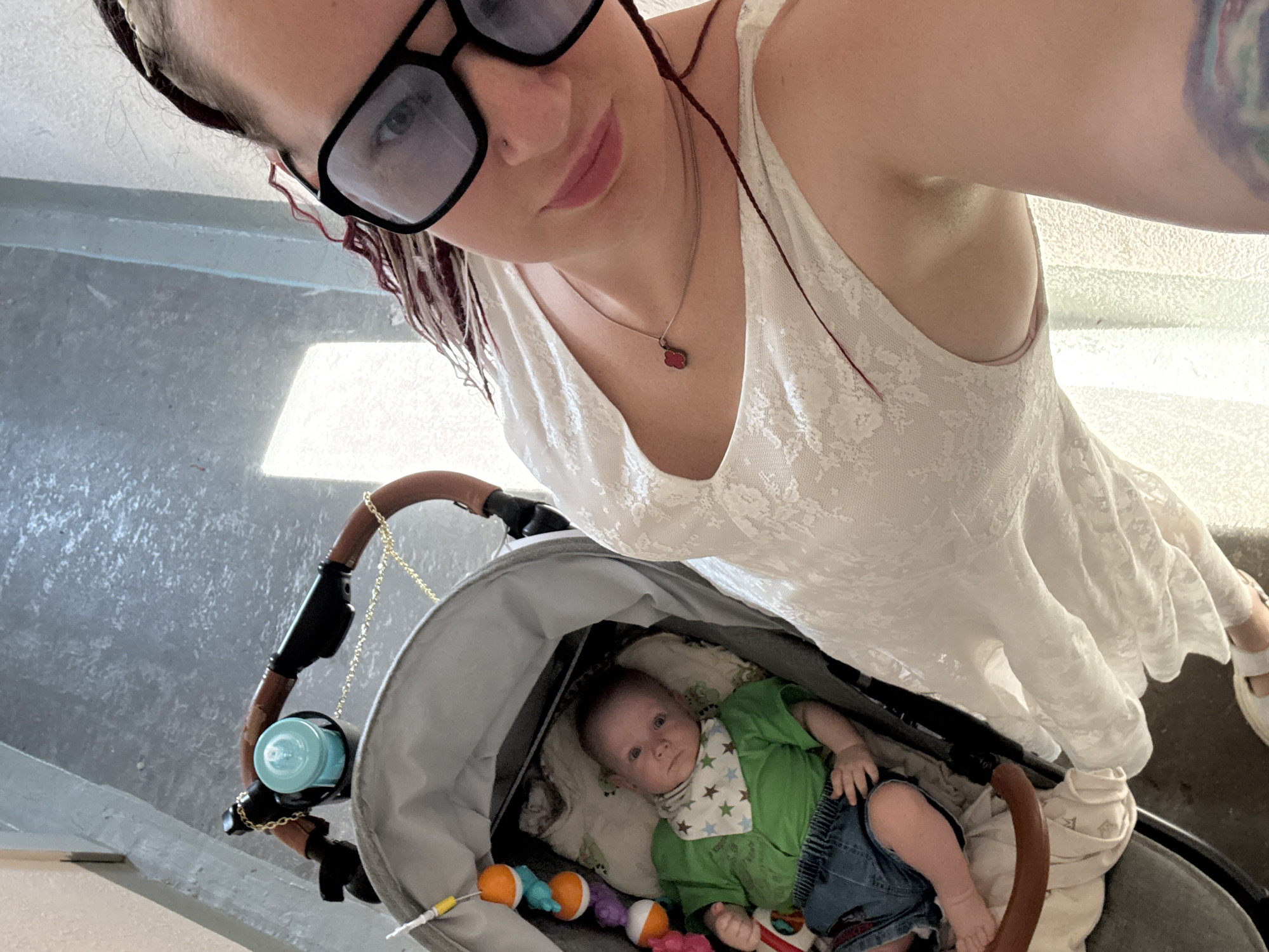 A high-angle selfie photograph captures an adult woman and an infant in a stroller within an indoor setting, described as taking place in Moscow, Russia.
The woman, positioned on the right side of the frame and angled slightly left, is looking directly at the camera. She wears a white lace sleeveless dress and black-framed sunglasses that partially obscure her eyes but reflect her right eye. A thin silver necklace with a small red four-leaf clover pendant is visible around her neck, and a colorful, abstract tattoo is on her upper left arm. Her lips are tinted pink, and her dark hair includes visible reddish-maroon strands.
To the woman's left, an infant lies in a light grey stroller, also looking towards the camera. The baby is dressed in a short-sleeved green shirt, denim-look shorts, and a white bib adorned with blue and green stars. A multi-colored mobile toy, featuring orange, blue, and purple elements, hangs above the baby. The stroller's handle is a dark brown, faux-leather material, accompanied by a decorative gold-colored chain and a cup holder containing a light blue baby bottle.
The background consists of a grey, textured floor and light-colored walls. Bright natural light enters the scene from the left background, creating a rectangular highlight on the floor.