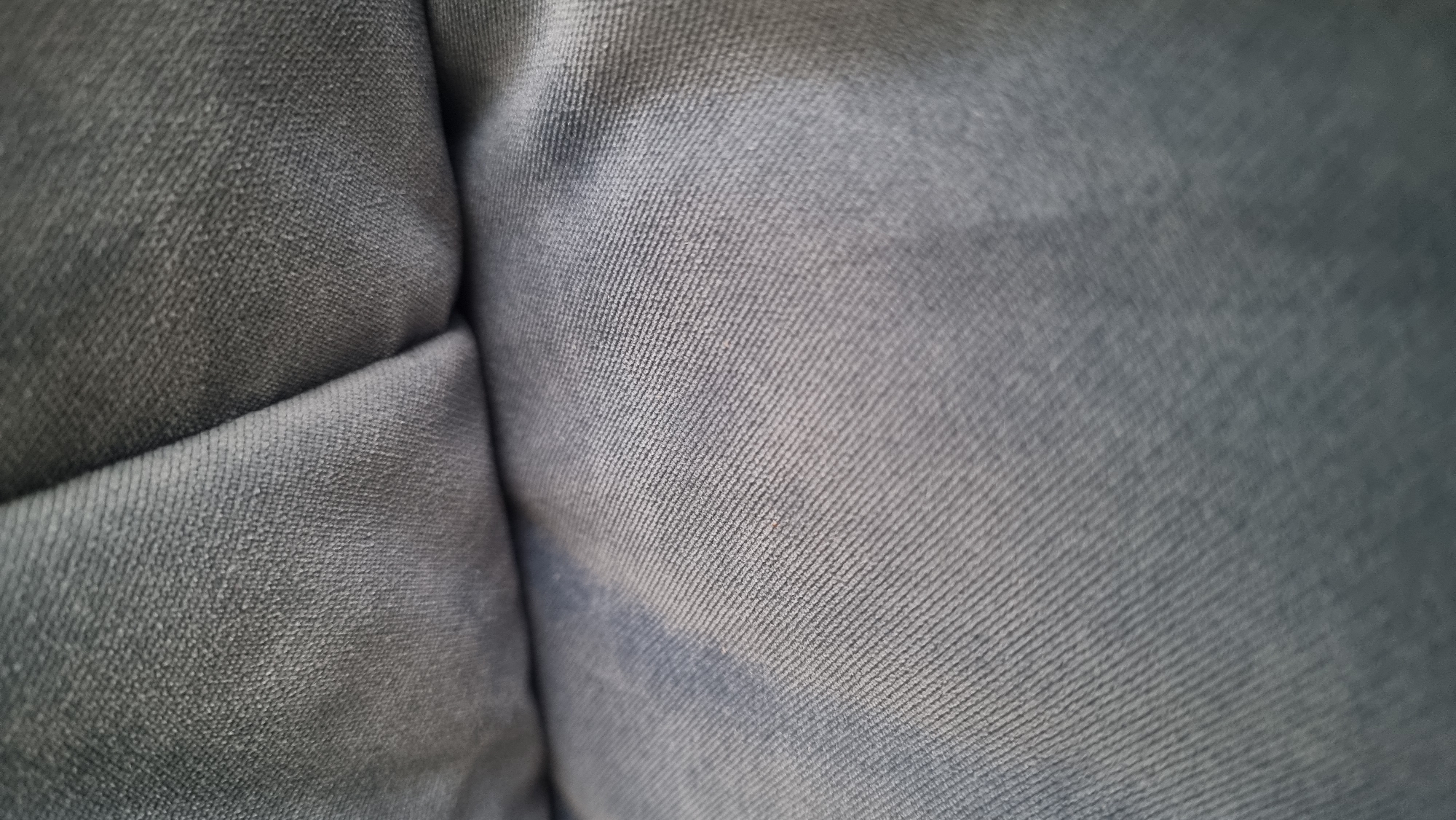 A close-up, top-down view of denim fabric is presented. The denim is a muted blue-grey color with visible diagonal lines characteristic of the twill weave. The fabric is creased and folded, creating shadowed areas and highlights that accentuate the texture. The lighting appears soft and diffused, with the light source coming from the upper left, casting subtle shadows and illuminating the raised threads of the denim. The image focuses solely on the textile, with no discernible background or environmental context. The scene is static and devoid of any human presence or activity.