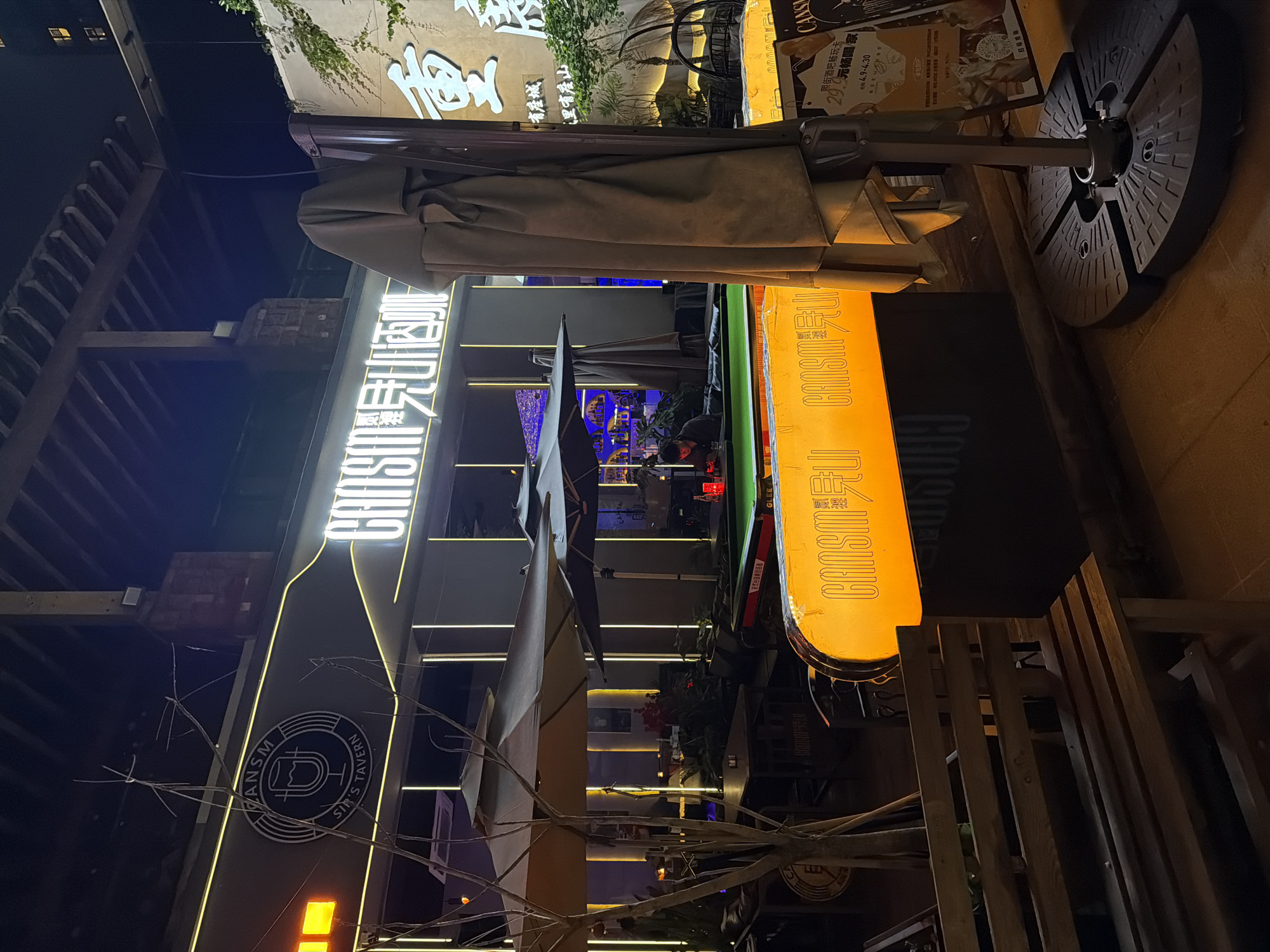 This nighttime image captures the vibrant outdoor area of a bar or tavern located in the Chongqing High-tech Zone, China. The scene is illuminated by numerous artificial lights, including prominent neon signs, casting a lively glow.
Key subjects include the exterior of the establishment, featuring several large umbrellas, one of which is prominently closed in the foreground. There are also outdoor seating arrangements, though mostly obscured. One person is partially visible in the mid-ground, seated at a table, suggesting patrons are present.
Notable details include the various glowing signs. A large, bottle-shaped sign on the left displays "CANSM SIR'S TAVERN" within a circular logo. Above it, a neon sign reads "CANSM 现山酒师". To the right, a large, rectangular yellow sign also says "CANSM". In the background, on a building facade with climbing greenery, large Chinese characters "重" are visible, possibly part of "重庆" (Chongqing) or a poetic phrase like "有座城 里有度山" ("There's a city, inside there's a mountain").
Promotional posters are affixed to a display on the right, advertising a "照街酒吧畅玩卡" (Zhàojiē Bar Play Card) for "29.9元畅阳家" (29.9 yuan unlimited drinks), valid from April 9th to April 30th, with a "扫码购" (scan to buy) option. The word "duke" and "GLEE" are also visible on other signs. The general atmosphere is that of an active entertainment venue in the evening.