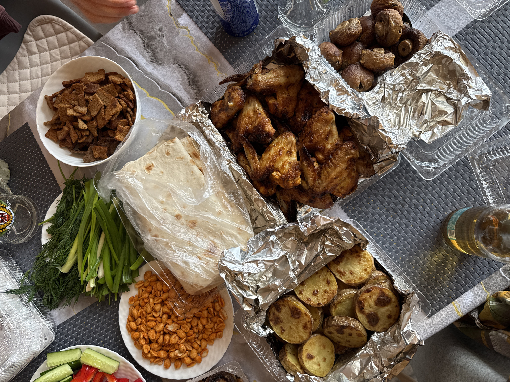 A top-down, eye-level, medium shot reveals a table laden with food, possibly outdoors or in a brightly lit room. The food items appear to be part of a meal or gathering. There are:
- A white bowl filled with croutons.
- A plate with green onions and dill.
- A plastic-wrapped package, likely containing flatbread.
- A plate with peanuts or similar legumes.
- A plate with sliced cucumbers and bell peppers.
- A disposable aluminum container with what looks like grilled or roasted chicken wings.
- Another aluminum container with roasted potatoes.
- A third aluminum container with mushrooms.
Other items include glassware and a beverage bottle. The location of the photograph is mentioned as Krasnoyarsk, Russia. There is an arm visible in the top left corner of the photograph. The table has placemats with woven design.