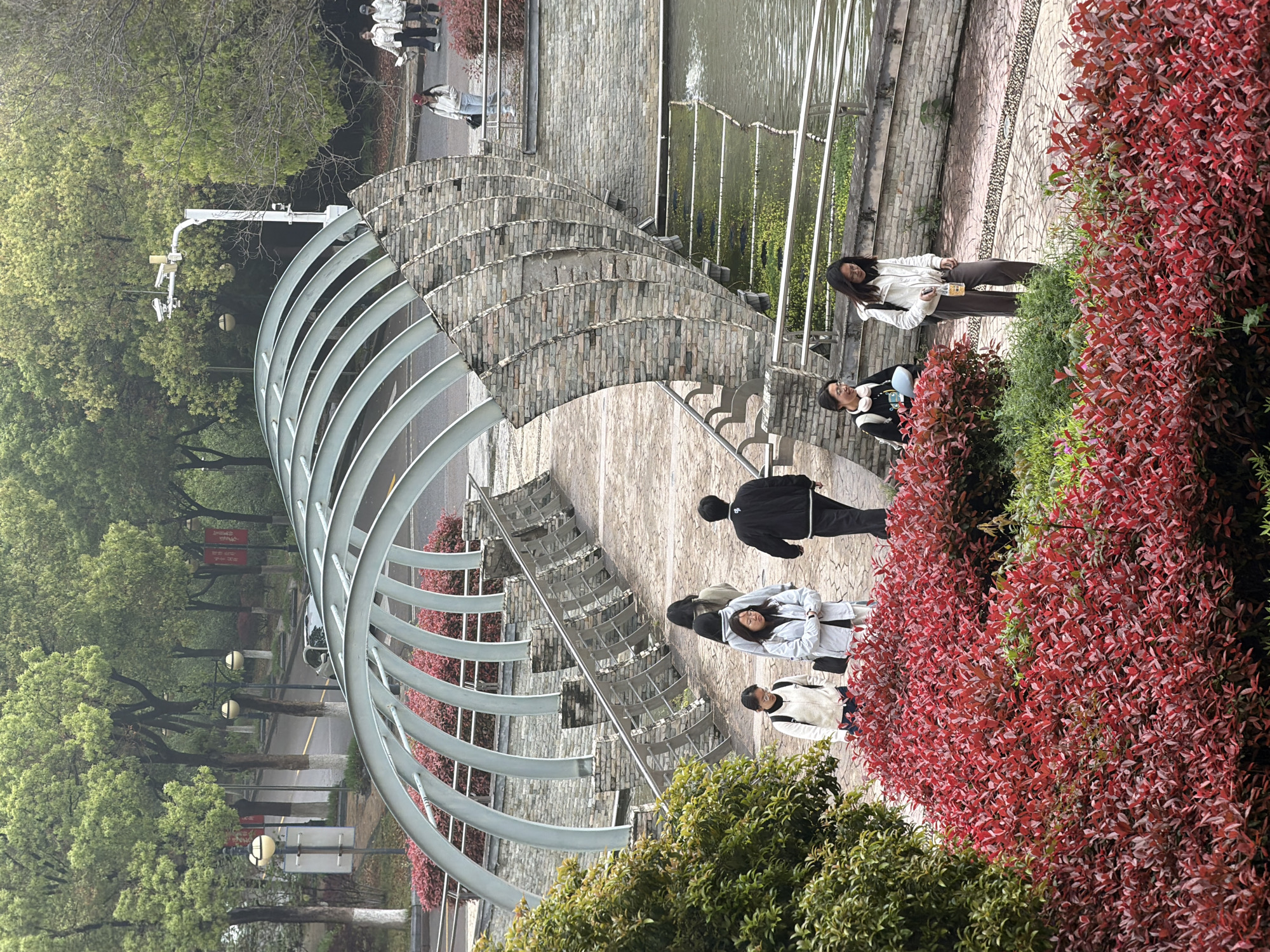 The image depicts a park scene in Jiangning District, China. A curved, architectural structure made of metal beams and stone brickwork spans a pedestrian walkway and a narrow waterway. Several individuals are present: One person walks away from the camera, wearing a black jacket; three individuals, dressed in light-colored outerwear, walk in the opposite direction; and two other people stand on the steps. Lush greenery, including trees and bushes with both green and red leaves, frames the walkway. In the background, a street runs parallel to the walkway, with lampposts and trees lining the side.