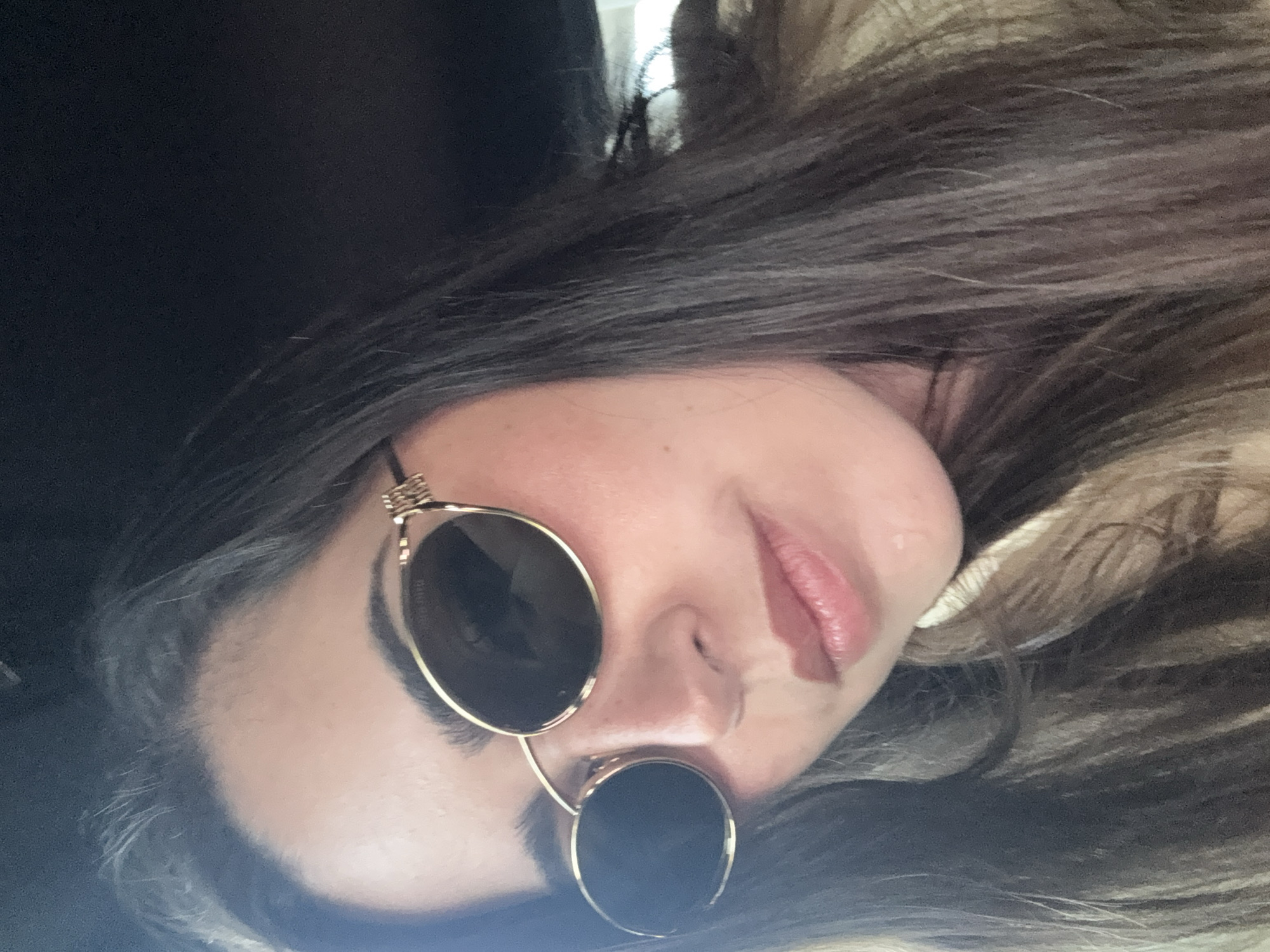 A woman is posing for a selfie. She is wearing sunglasses with round lenses and gold frames. Her hair is long and brown. The background is blurry, but it appears to be a dark interior. The location of this image is unknown, and it is not possible to determine if it was taken in Mexico City.