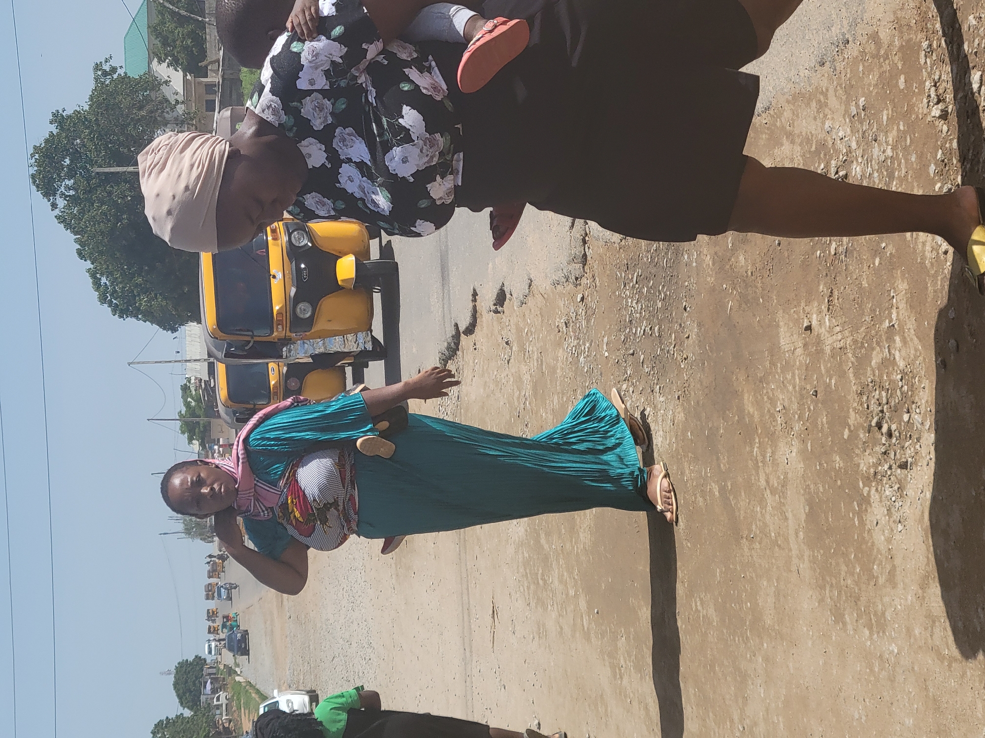 In Kwamba, Nigeria, on a sun-drenched day, two women walk along a dusty, unpaved road. The woman on the left, wearing a turquoise pleated dress and a pink and patterned scarf, carries a baby wrapped in a colorful cloth across her chest. Her head is bowed slightly, and her expression is neutral. The woman on the right, wearing a black floral-patterned top and a dark skirt, has a baby on her back, secured with a fabric wrap. She looks down, and her expression is also unreadable. In the background, several yellow auto-rickshaws, known locally as "keke napep," are driving on a paved road. The environment is characterized by sparse vegetation, scattered buildings, and utility poles, indicating a semi-urban or developing area. The overall impression is one of everyday life in a Nigerian town, with people navigating their daily routines.