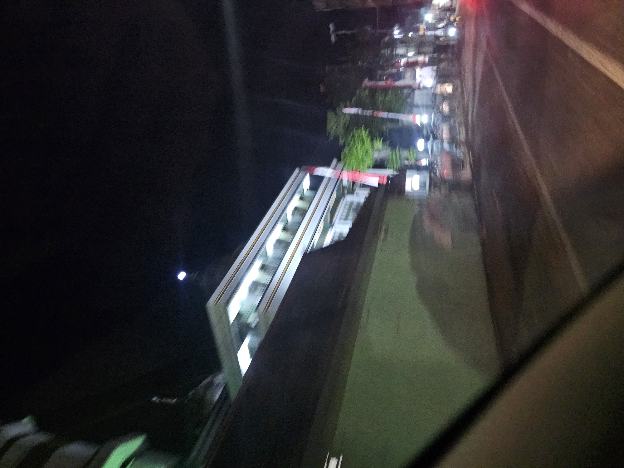 The image is a night-time, blurry photograph taken from inside a vehicle, likely in Jenu, Indonesia. The scene captures a street view featuring a building with multiple levels, illuminated by interior lights. In front of the building are several tall, red and white flags, which appear to be Indonesian flags. Trees are visible behind the flags. The street itself is paved with white lane markings, and there are lights from other vehicles and possibly streetlights. The overall impression is one of a busy urban environment at night.
