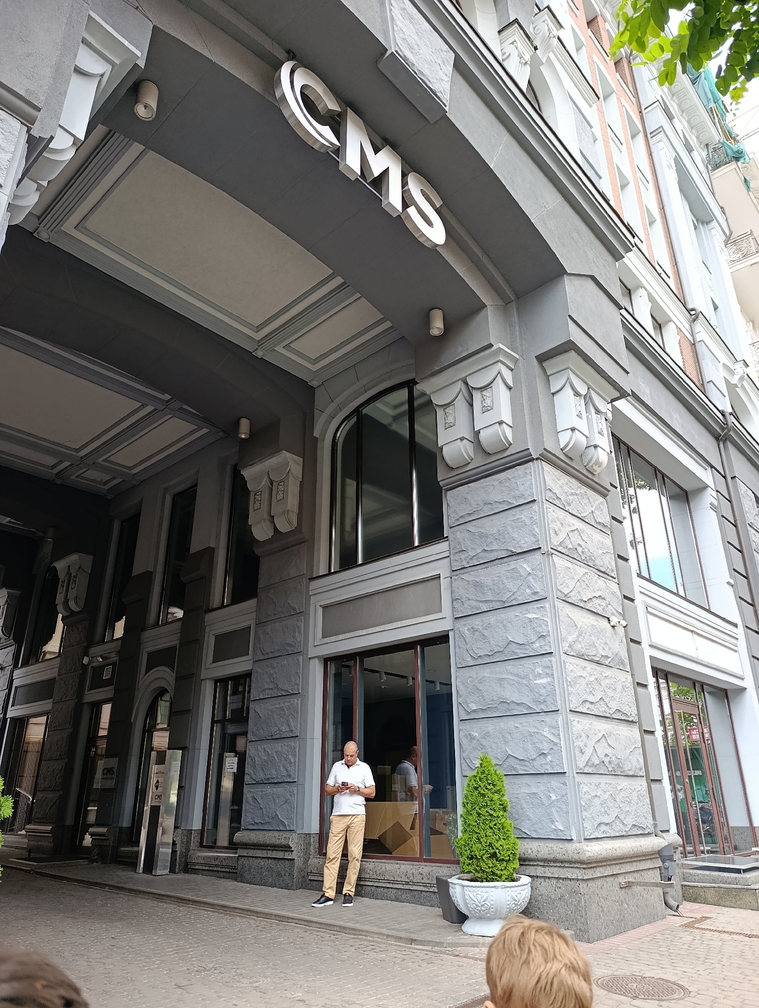 A man in a white polo shirt and tan pants stands on a sidewalk looking at his phone. He is standing outside of a modern building with large windows. The building has a sign with the letters "CMS" on it. The man is bald and appears to be middle-aged. The building is in Kyiv, Ukraine. The sidewalk is paved with cobblestones. There is a small evergreen tree in a white pot next to the man.