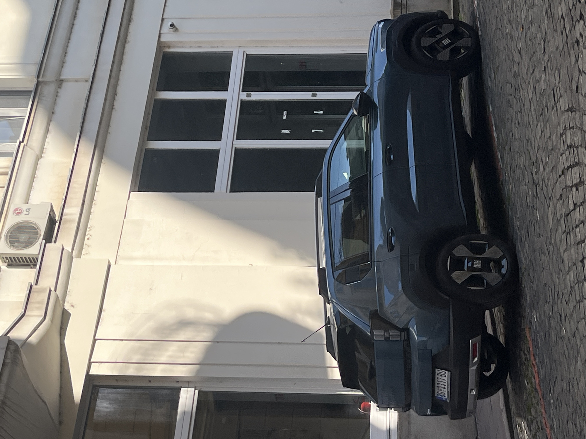 A dark blue Fiat 600 Electric is parked on a cobblestone street in front of a light-colored building. The building has several large windows, some of which are covered with dark film, and an air conditioning unit is visible near the top. A shadow from a tree or structure is cast on the side of the building. The car has Serbian license plates that read "BG 3317-BU". The street is paved with cobblestones, and a thin red line is visible on the ground in front of the car. The image is taken from a slightly elevated side-angle, focusing on the rear and side profile of the vehicle.