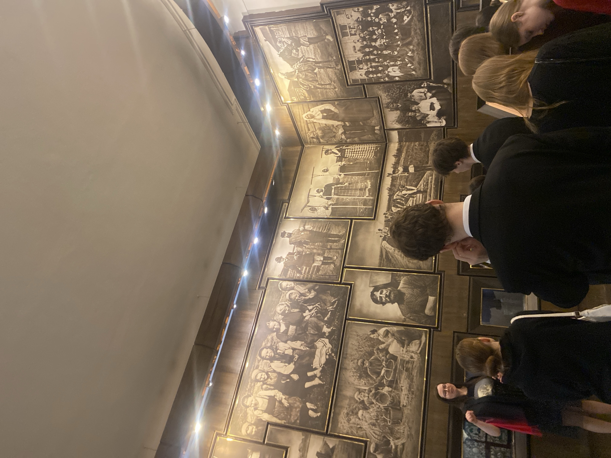 A group of people are gathered in what appears to be a museum or gallery in Saint Petersburg, Russia, looking at a wall of framed black and white historical photographs. The photos vary in subject matter, including portraits of people, groups of people, and outdoor scenes with figures engaged in activities. The people observing the photos are dressed in dark-colored clothing, primarily black, suggesting a formal or somber occasion. The setting includes architectural details like a curved ceiling and lighting fixtures, suggesting an interior space designed for display.