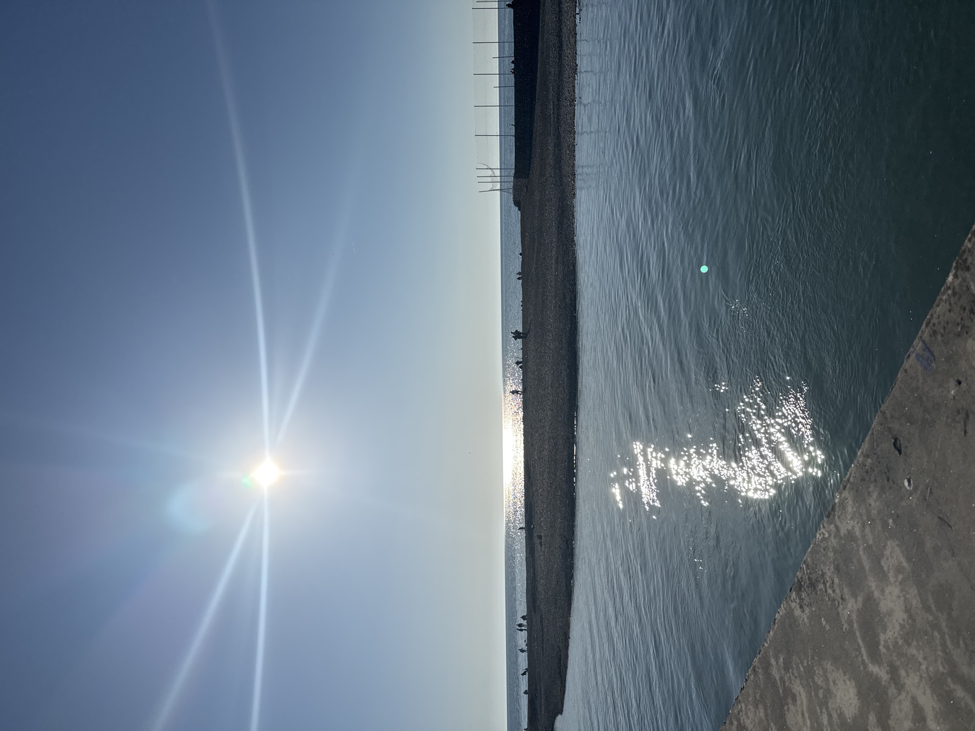 A landscape-oriented photograph depicts a sunny, outdoor scene with a horizon line running vertically through the center. To the left, the sky is bright blue with the sun positioned near the bottom. Sunlight streams upward. To the right, the ocean appears textured. The horizon line reflects sunlight. The scene appears to be on a beach with a fenced off section, with people standing on the gravel. The location is Unknown, Unknown.