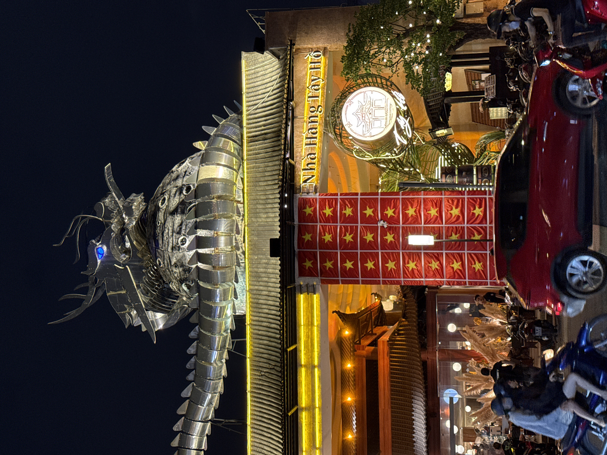 Captured at night, the image showcases the "Nhà Hàng Tây Hồ" restaurant in Thủ Đức, Vietnam. A large metallic dragon sculpture, illuminated with a blue eye, stands to the left of the building. The restaurant features a sign with neon lettering and is decorated with Vietnamese flags. A parked red car and several motorcycles are visible in the foreground. Pedestrians are present near the entrance of the restaurant.