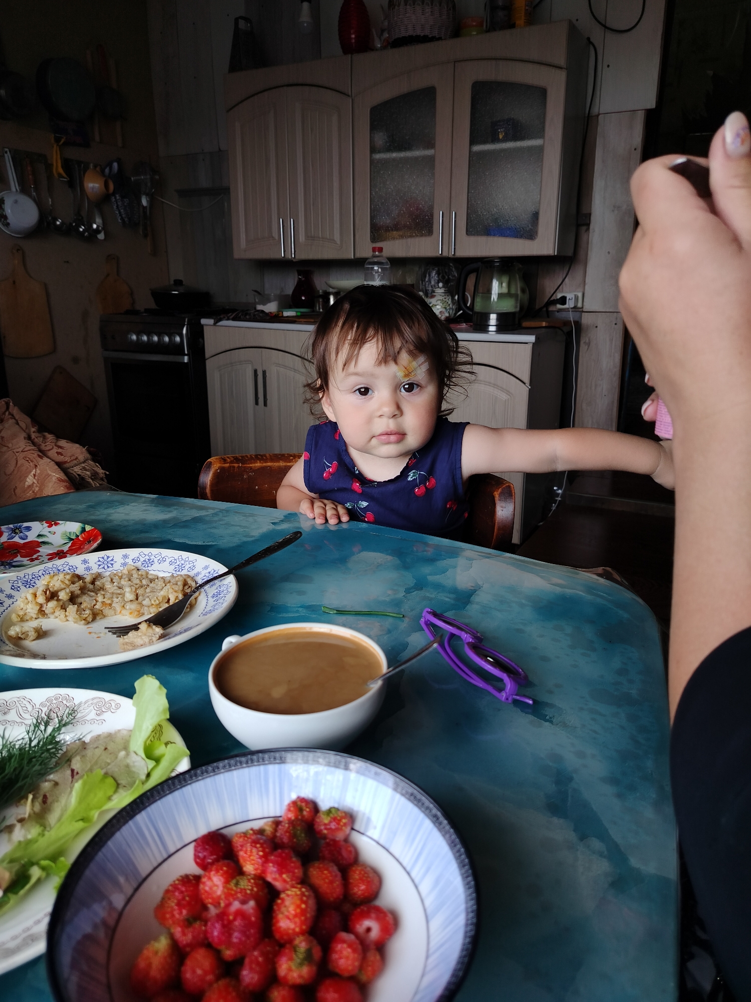 In a dimly lit kitchen, a young child sits at a turquoise table. The child, wearing a dark blue dress with cherry patterns and sporting a bandage on their forehead, gazes directly at the camera. In front of the child is a plate of food and a bowl of strawberries. A person's hand, with painted nails, is visible in the foreground, reaching towards the child. The kitchen environment shows cabinets, a stove, and various kitchenware, suggesting a domestic setting in the city of Блохино, Russia.