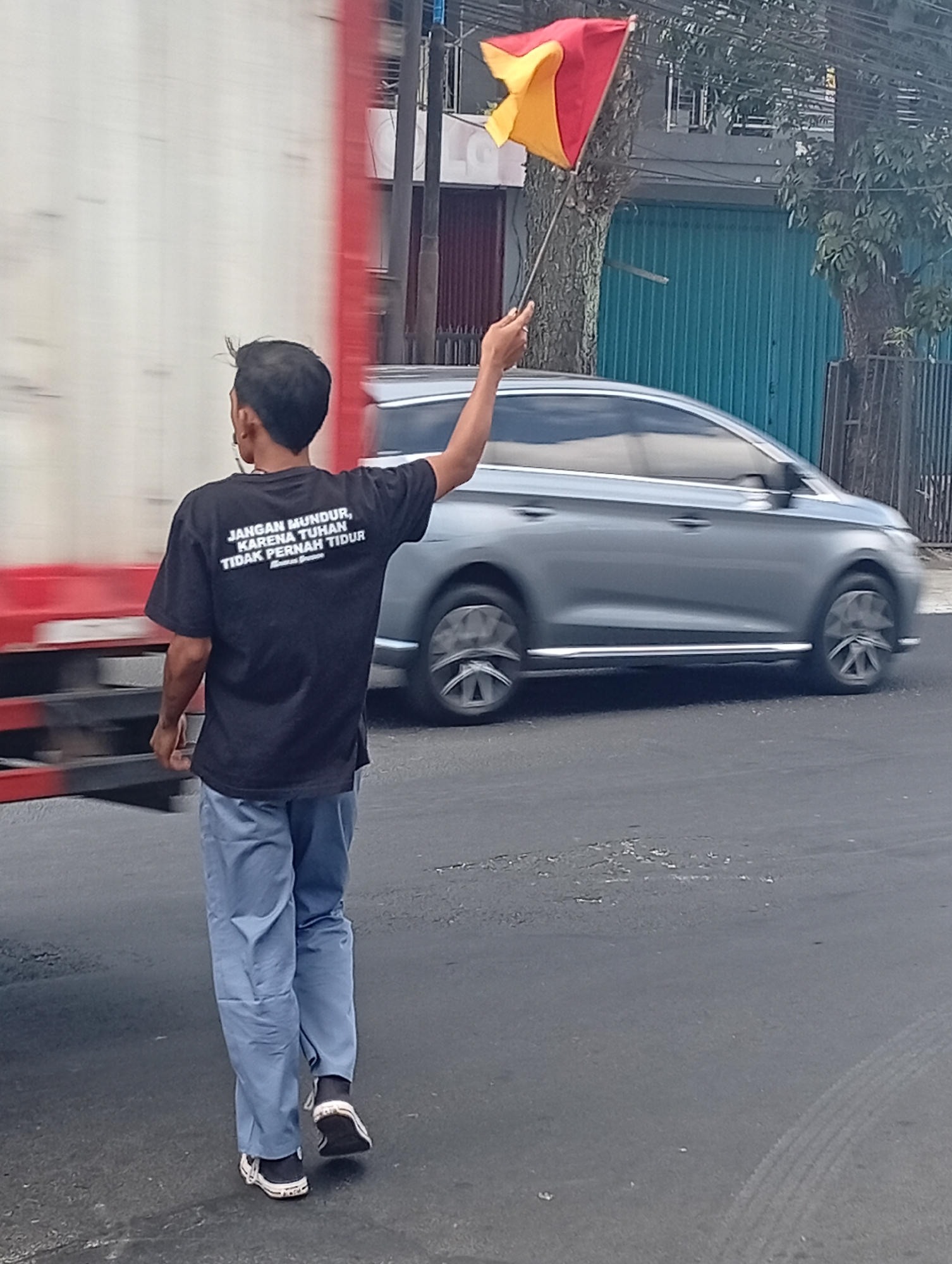 A person wearing a black t-shirt and light blue pants is standing on a paved road, holding a red and yellow flag. The t-shirt has white text on the back that reads "JANGAN MUNDUR, KARENA TUHAN TIDAK PERNAH TIDUR" which translates to "Don't retreat, because God never sleeps". A large truck, predominantly white with a red stripe, is visible to the left. A silver car is driving past on the right, with motion blur indicating movement. In the background, there are buildings with teal and brown facades, trees, and power lines. The scene appears to be on a street in Malang, Indonesia, based on the text on the shirt and the style of the surrounding environment.