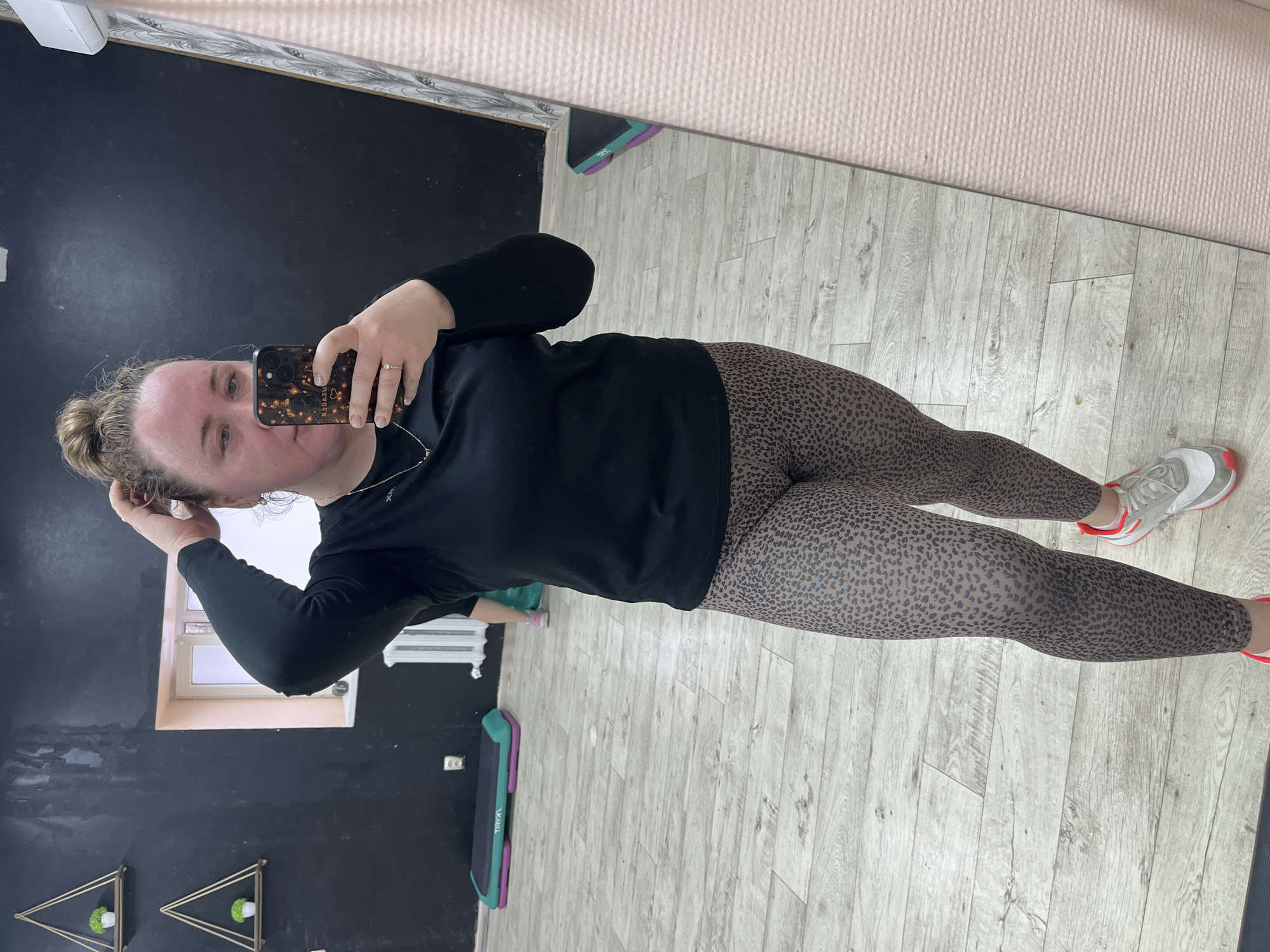 A medium-shot, low-angle image captures a woman in a fitness studio setting. She is holding a smartphone to take a selfie in a mirror. The woman is wearing a long-sleeved black shirt and animal-print leggings. Her footwear consists of athletic shoes with red accents.
The background features a black wall and a wooden floor. Elements such as a window, hanging plant decorations, and exercise step equipment are present. The location is potentially in Chaikovsky, Russia.