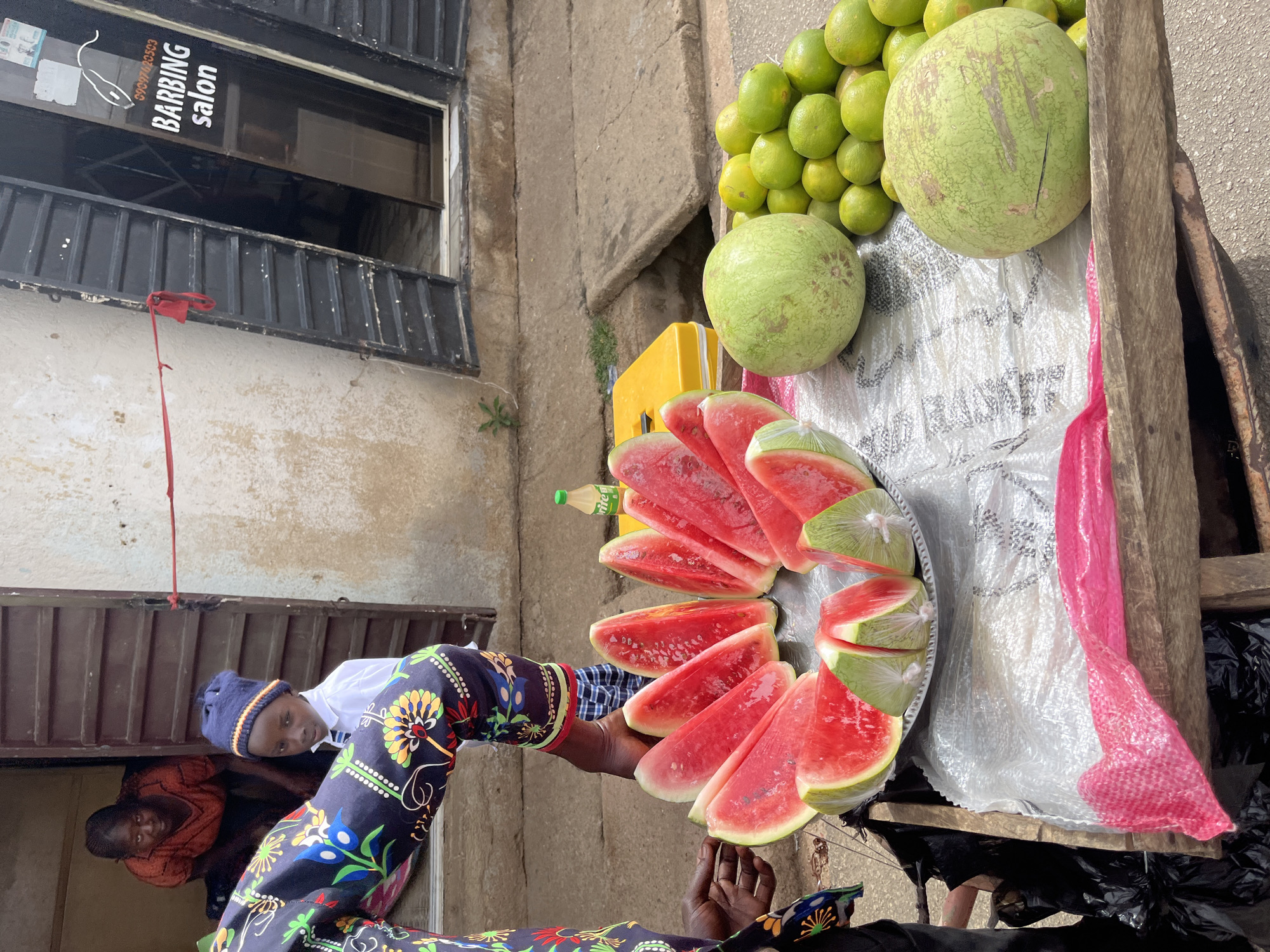 In Barakin Akawo, Nigeria, a vendor displays watermelons and green fruits on a wooden cart. Sliced watermelons are arranged on a platter. A boy wearing a blue hat and patterned pants is partially visible. A "BARBING salon" sign is visible in the background. Another person in orange clothing is partially visible below the boy. A yellow container sits among the watermelons.