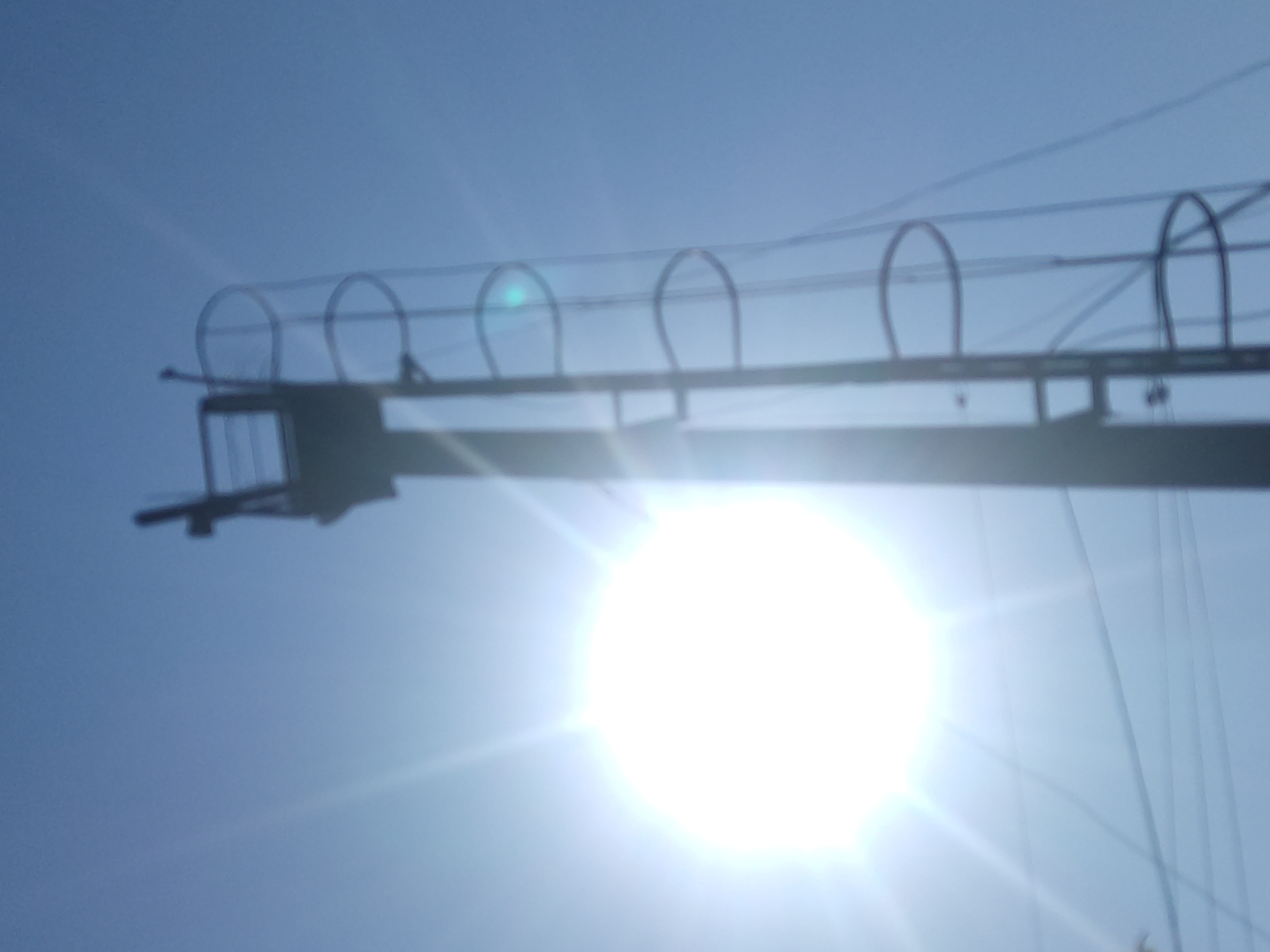 The image captures a bright, sunlit sky with a tall, dark structure, possibly a transmission tower or lighthouse, dominating the right side of the frame. The structure has a ladder-like ascent with circular protective cages at intervals. The sun is prominently positioned in the left half of the image, emitting strong rays that cause lens flare and overexpose a portion of the sky. Several thin, dark lines, likely power lines, are visible in the lower part of the image. The overall scene suggests an outdoor, daytime setting. There is no specific information to indicate the location within the city of Radisne, Ukraine.