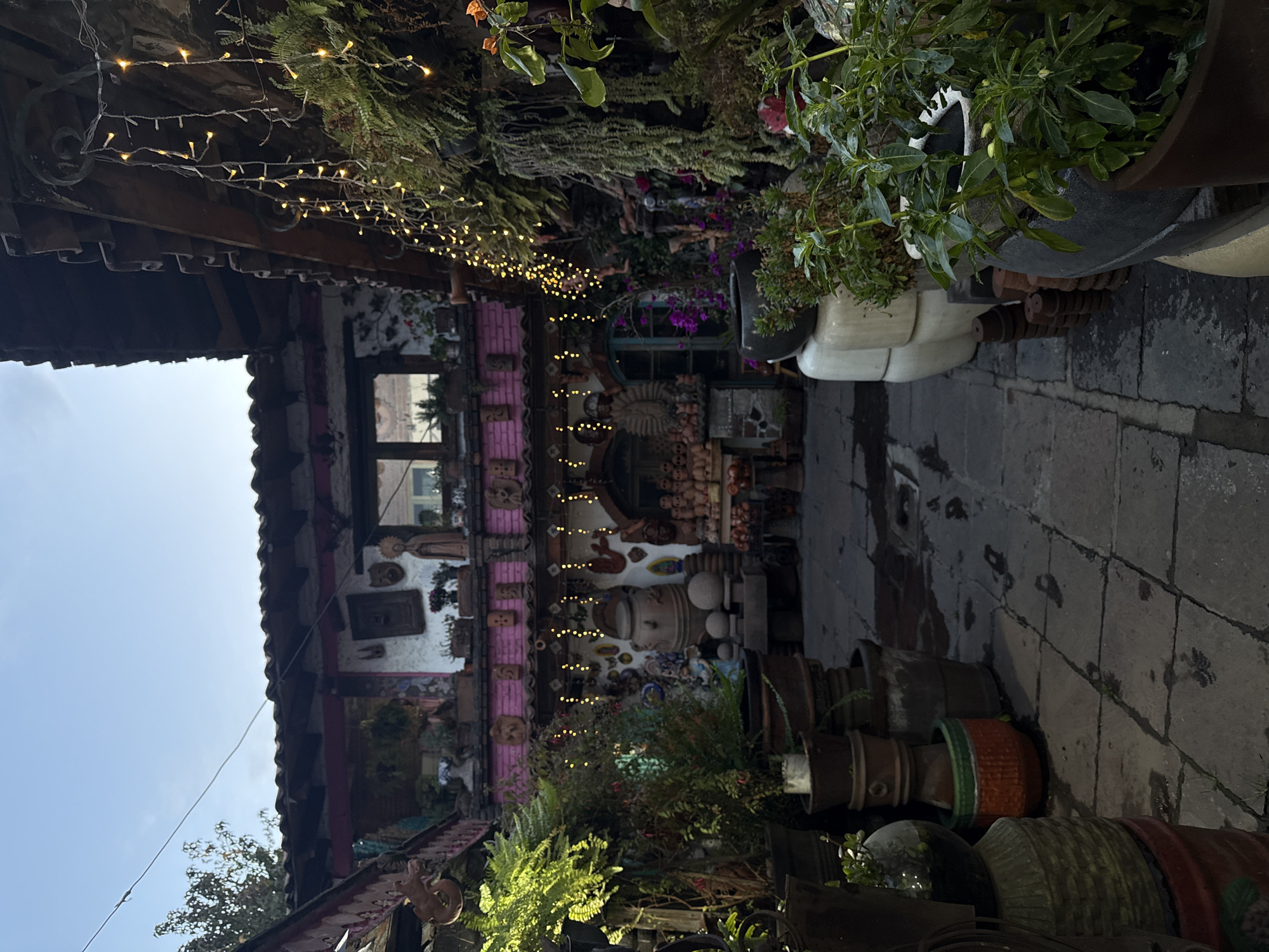 This outdoor scene appears to be a courtyard or pathway within a building complex located in Metepec, Mexico. The architecture includes a building with a pink and white facade, adorned with various decorative items, sculptures, and pottery. Strings of lights are strung across the top, adding a festive atmosphere. The foreground is filled with an assortment of potted plants, clay pots, and other decorative objects. The ground is paved with stone tiles. There are no people visible in the scene. The overall impression is of a creatively decorated and well-maintained space.