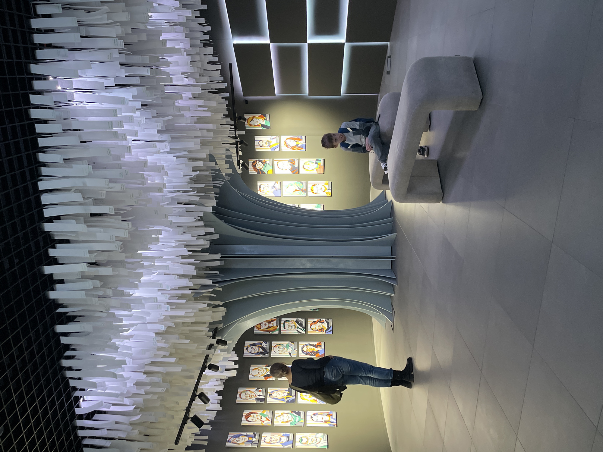 A person stands in an interior space, presumably a gallery or museum. The space features modern design elements including a black grid ceiling with hanging white rectangular shapes that form a cloud-like installation, and a curved architectural structure displaying a series of framed portrait artworks on the wall. The person is looking at the art. In the background, another person is seated on a curved gray sofa. Overhead lighting illuminates the ceiling and artwork, and a light tile floor complements the modern aesthetic. The space is located in Moscow, Russia.