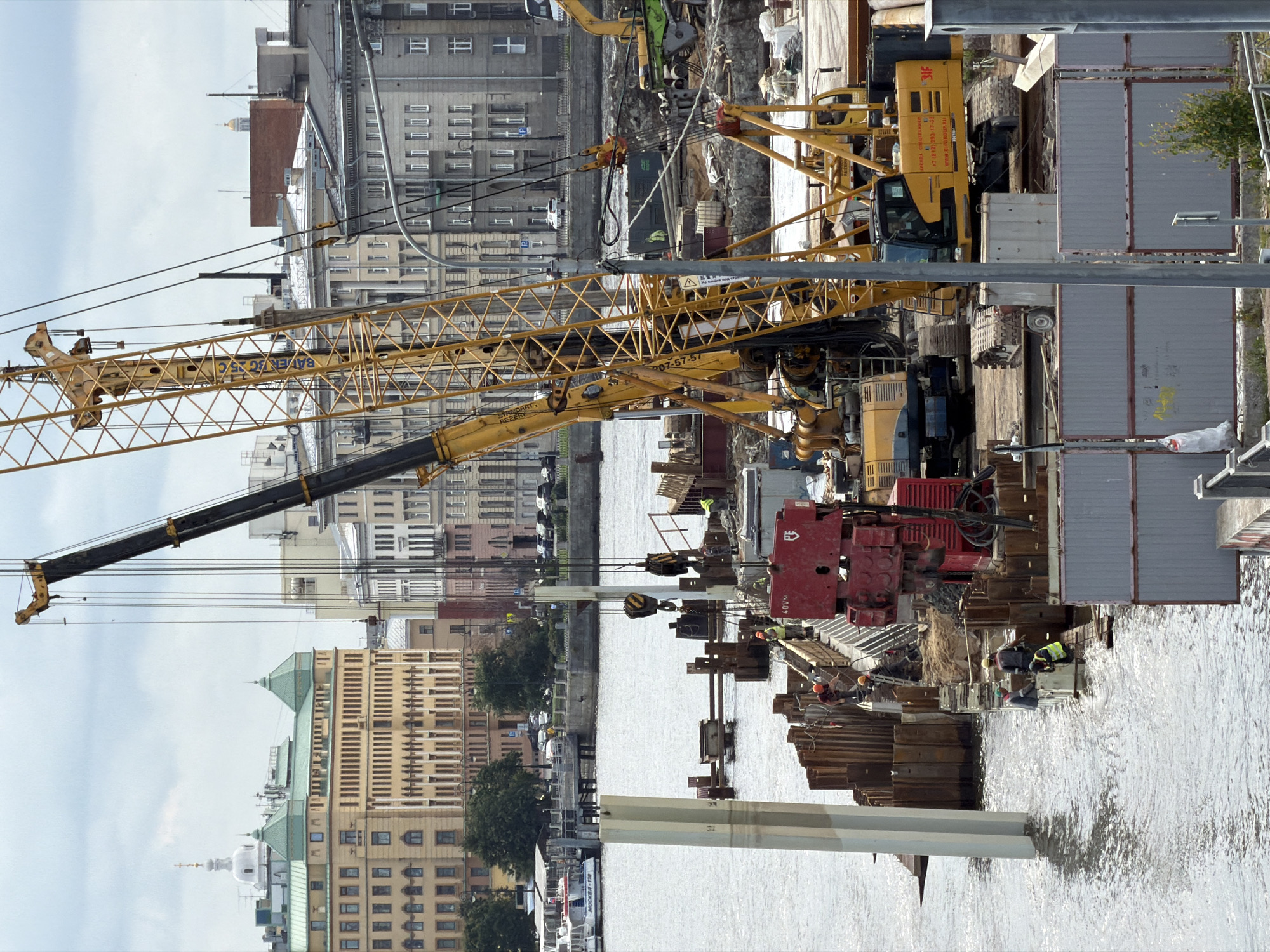 The image shows a construction site on the Neva River embankment in Saint Petersburg, Russia. A large yellow crane is prominently positioned, with its boom extending upwards. In the foreground, there is a partially submerged concrete pillar and a sheet pile wall, indicating ongoing construction work to reinforce the embankment. Several workers, wearing hard hats and safety vests, are visible on the construction platform, engaged in various tasks. Construction equipment, including smaller yellow excavators and a red machine with the inscription "40VN", are also present. In the background, the architectural cityscape of Saint Petersburg is visible, featuring historical buildings with distinctive green roofs and a white dome. The Neva River flows in the foreground, with a few boats docked on the far bank. The sky is overcast.
