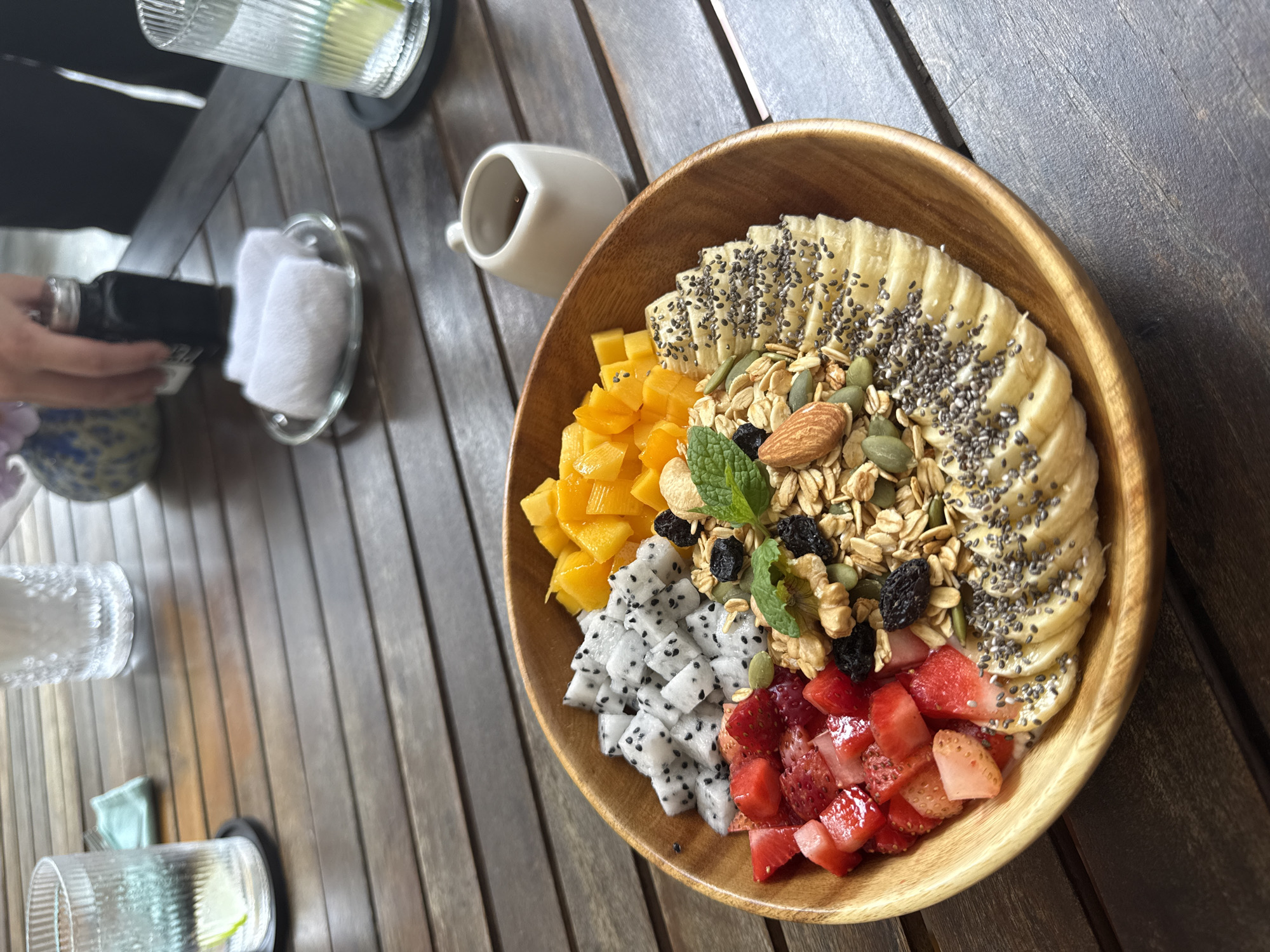 A wooden bowl filled with a colorful assortment of fruits and granola is centered in the frame. The fruits include sliced bananas topped with chia seeds, diced mango, cubes of dragon fruit, and diced strawberries. A mix of granola, nuts, and seeds occupies the spaces between the fruits. A sprig of mint is nestled among the granola. The bowl sits on a wooden table with a slatted pattern.
In the background, a small white pitcher is visible, along with a clear glass of liquid with a lime wedge. Other glasses and a hand holding a black device are partially visible on the left. A rolled white towel is placed on a small glass dish in the upper left corner. The scene suggests a cafe or restaurant setting.
While specific location details are not explicitly provided in the image, the query requests the city of Đà Nẵng, Vietnam. Given the food presentation and restaurant-like setting, it is plausible the image was taken at a cafe or eatery in Đà Nẵng, Vietnam, serving a breakfast or brunch dish.