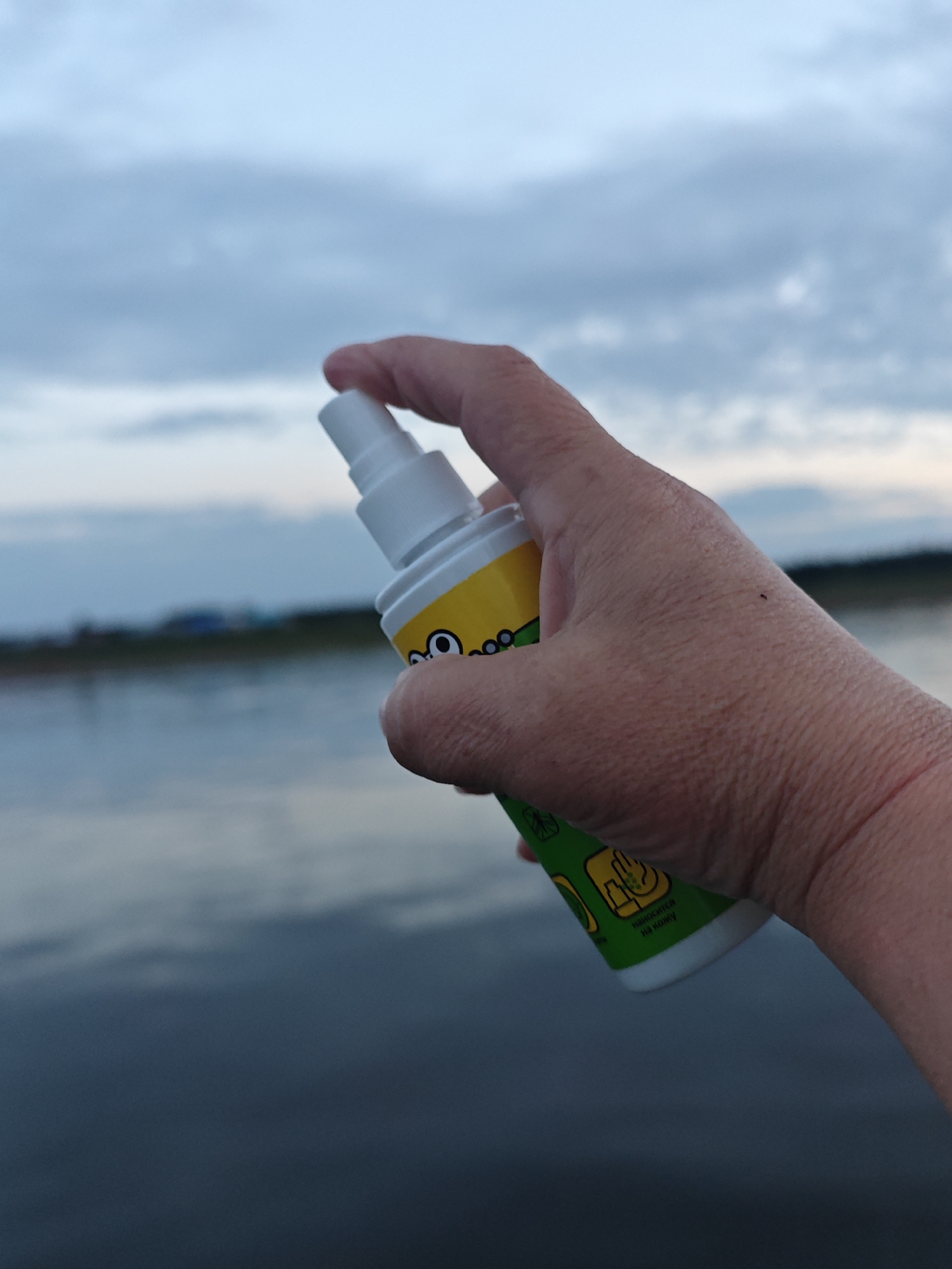 A close-up shot shows a person's hand holding a white and yellow spray bottle. The hand is positioned in the foreground, with the spray bottle angled slightly away from the camera. The bottle has a white cap and a yellow body with cartoon-like graphics. The lower part of the bottle is green with some text.
The background features a body of water, likely a lake or river, under a cloudy sky. The water is a deep blue-grey, reflecting the overcast conditions. In the distance, a faint line of trees or land is visible, with what appear to be small structures. The sky is a mix of grey and pale blue, suggesting either early morning or late evening. The lighting is diffused, consistent with cloudy weather.
The person appears to be outdoors, possibly near a body of water. The image conveys a sense of calm or routine activity, such as applying insect repellent before engaging in outdoor activities.
The visible text on the green part of the bottle is in Cyrillic and reads "Наносится на кожу," which translates to "Applied to skin." This suggests the product is a topical spray, likely for protection from insects or as a skin treatment. The overall impression is one of a practical moment captured in an outdoor setting.