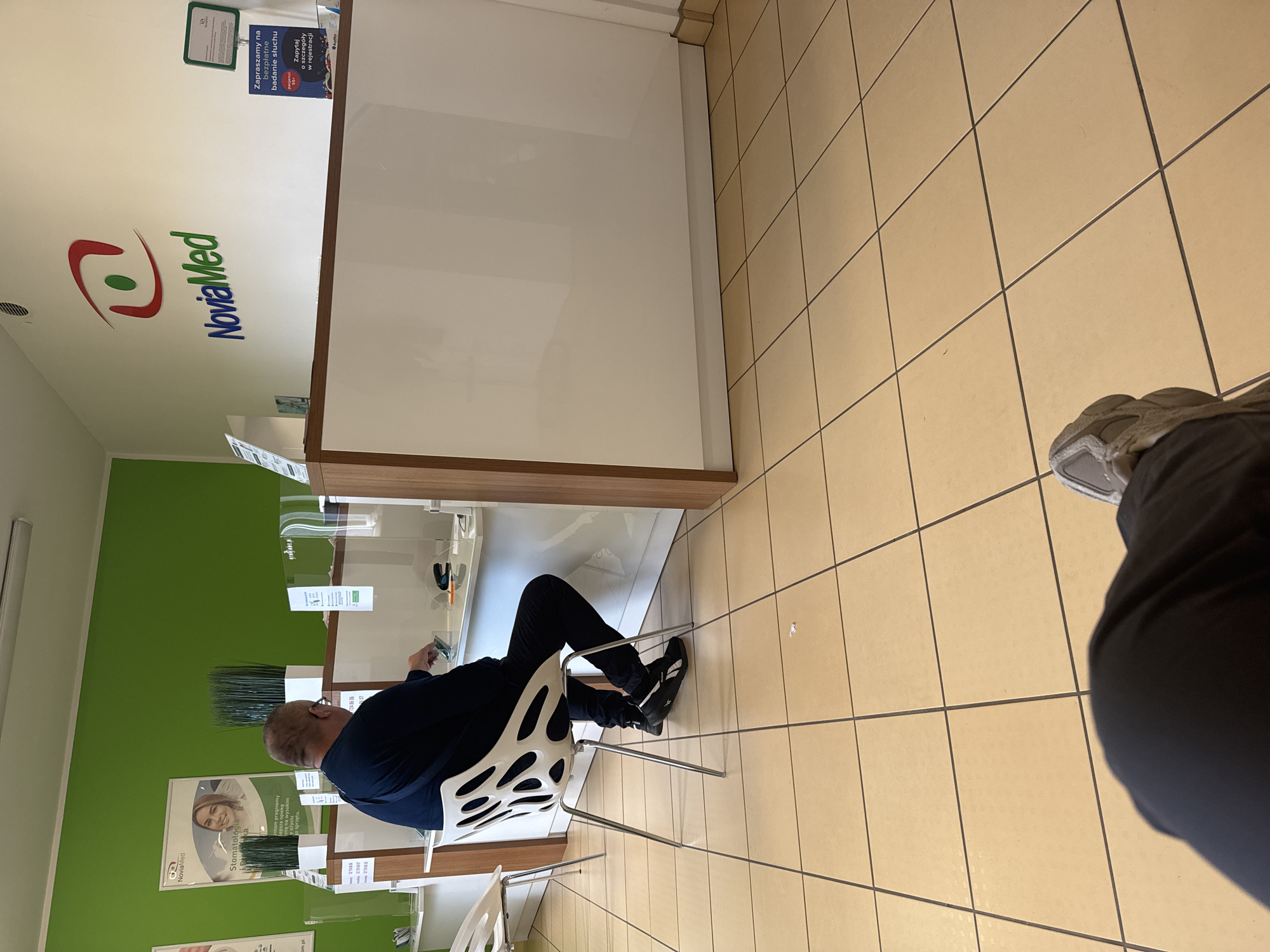 The image depicts the interior of a clinic or reception area. In the foreground, from a seated perspective, the legs and feet of an individual are visible, wearing dark pants and grey sneakers. The floor is tiled with large, light beige square tiles.
In the mid-ground, a man is seated on a modern white chair with a distinctive organic design. He is wearing a dark blue long-sleeved shirt and dark trousers, with black sneakers. He is facing a reception counter, leaning slightly forward and looking down at his hands, which are holding what appears to be a small white object. The reception counter is a two-tiered structure with a white glossy top surface and wooden trim. Partitions made of clear plastic are visible on the counter, separating different service points.
In the background, a vibrant green wall dominates the upper portion of the scene. On the wall, a logo and name "NoviaMed" are prominently displayed in red, green, and blue lettering. There are also posters and informational signs on the wall and on the counter, including one that reads "Zapraszamy na bezpłatne badanie słuchu" (We invite you for a free hearing test). A white bin is positioned near the counter. The overall environment appears clean and modern, suggesting a medical or healthcare facility. The signage, including the company name "NoviaMed" and Polish language text, indicates the location is likely in Poland. Given the context of medical services advertised and the general appearance, it is plausible this is a branch of NoviaMed in Chorzów, Poland, known for providing various medical services.