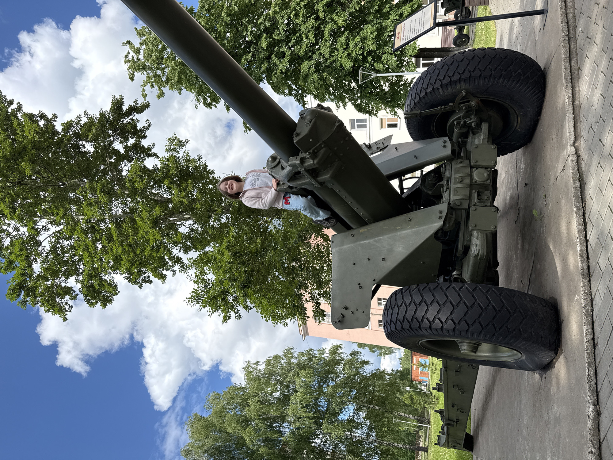 The image is a medium shot of a young girl sitting on a large, green artillery cannon in a public setting. The cannon has a long barrel pointing upward and is mounted on a wheeled chassis. The girl is wearing a light pink jacket, a gray t-shirt, and blue jeans. She is smiling and appears to be posing for the photograph.
The background includes trees, a blue sky with clouds, and a building. A sign is visible near the cannon, suggesting that it is part of an outdoor exhibit. Based on the presence of the M-46 cannon, the image appears to be taken at the Kalashnikov museum in Izhevsk, Russia.