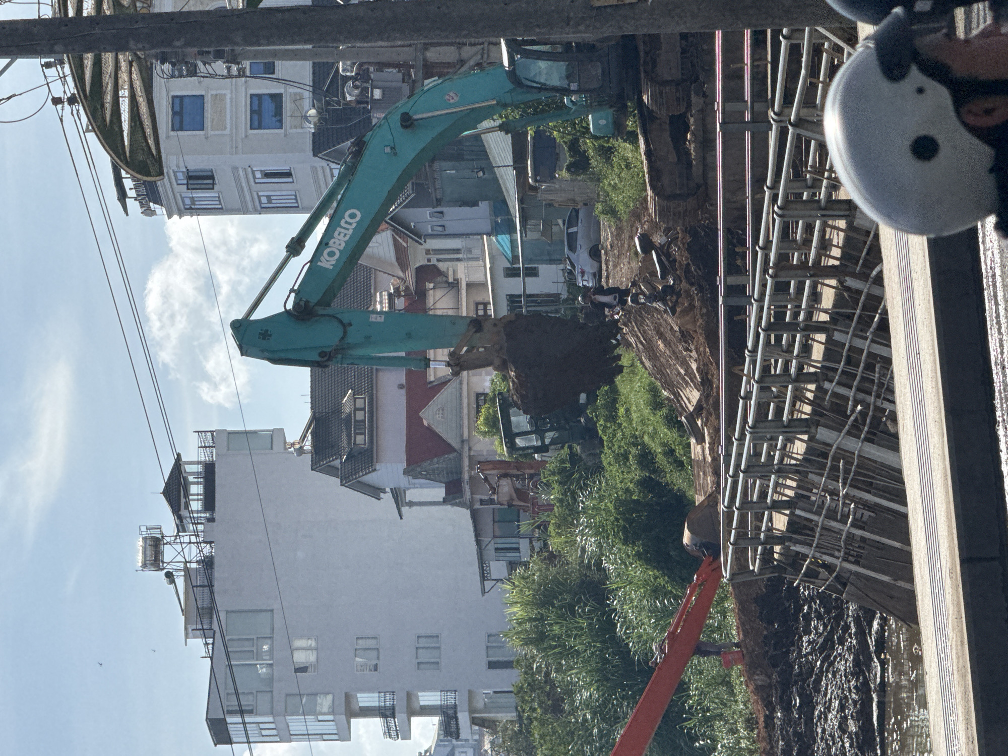 This detailed image captures an active excavation site adjacent to a muddy waterway in Da Lat, Vietnam, under a bright, clear sky.
In the midground, a large teal Kobelco excavator is actively digging into a mound of dark brown soil, its bucket full. To its immediate left, another older, green excavator is partially visible amidst dense green vegetation. Further left and lower, a red excavator arm extends towards the muddy water. A silver sedan is parked on a dirt path behind the teal excavator, and a black motorcycle is parked on the soil mound near the base of the excavator. Several workers are visible on and around the soil mound; one worker in a white shirt and dark pants stands observing the teal excavator's operation.
The foreground features a partial view of a person wearing a white motorcycle helmet with black dots, indicating they are on a motorcycle or scooter on a paved surface, likely a road or bridge. Behind this person, a concrete structure with a metal railing, possibly a bridge or retaining wall, spans horizontally. Sections of this railing appear to be under repair or damaged, with exposed rebar or wooden supports extending into the murky, muddy water below. The water surface shows significant dark mud accumulation, suggesting recent earth displacement or a high water level.
The background is dominated by various buildings and lush green hillsides. On the left, a modern, multi-story white building with extensive glass windows and balconies features a rooftop structure with a solar water heater and a small cupola. In the center-right background, another multi-story white building with blue-tinted windows and decorative cornices has a partially visible sign on its roof, reading "AN BAO." Numerous traditional Vietnamese houses with dark grey and red tiled roofs, some with gables and verandas, are interspersed among the larger structures. Overhead, multiple power lines and utility poles are visible, traversing the scene.