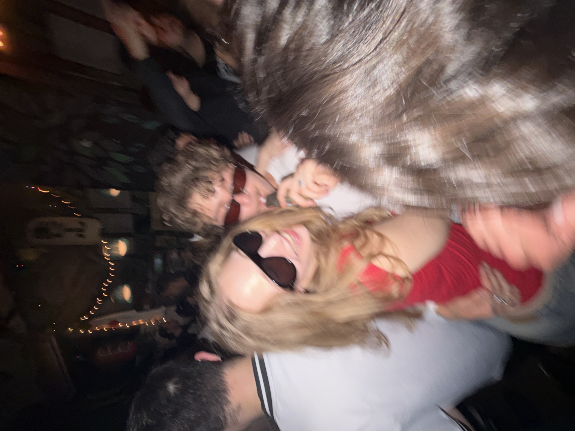 The image is a motion-blurred photograph taken indoors, likely at night in a dimly lit venue with string lights visible in the background. In the foreground, three individuals are partially visible. On the left, a person wearing a white t-shirt with a black collar is seen from the back and side. In the center, a young woman with blonde hair is smiling and wearing large, dark sunglasses and a red top. She is holding a dark object to her mouth. To her right, a young man with curly hair and red-tinted sunglasses is also visible. The overall atmosphere suggests a social gathering or party. The specific location in Belgrade, Serbia, cannot be determined from the image alone.